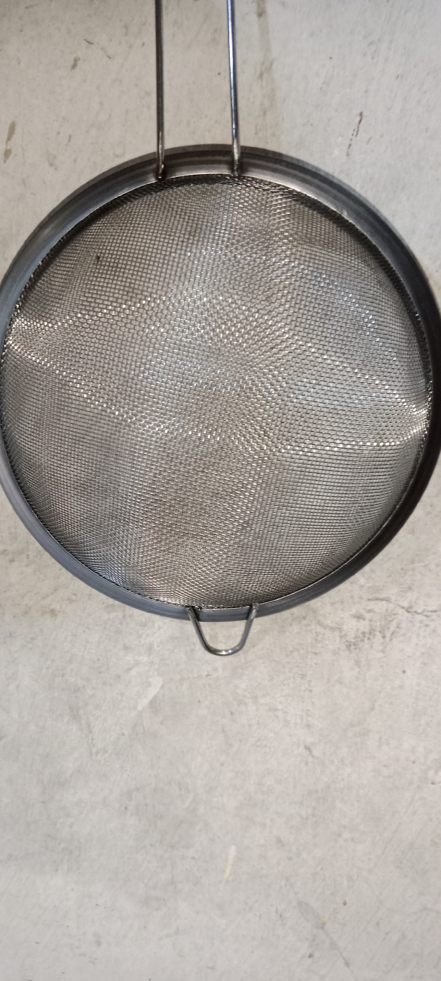 A stainless steel mesh sieve with two handles is suspended from above, casting a shadow on a light gray concrete wall. The sieve has a wide, flat rim and a fine mesh interior. The concrete wall appears to be the exterior of a building, possibly in an urban environment. The lighting suggests it is daytime, with natural light illuminating the scene. The image does not contain any identifiable landmarks or features that would specifically place it in Subang, Indonesia, or any other city.