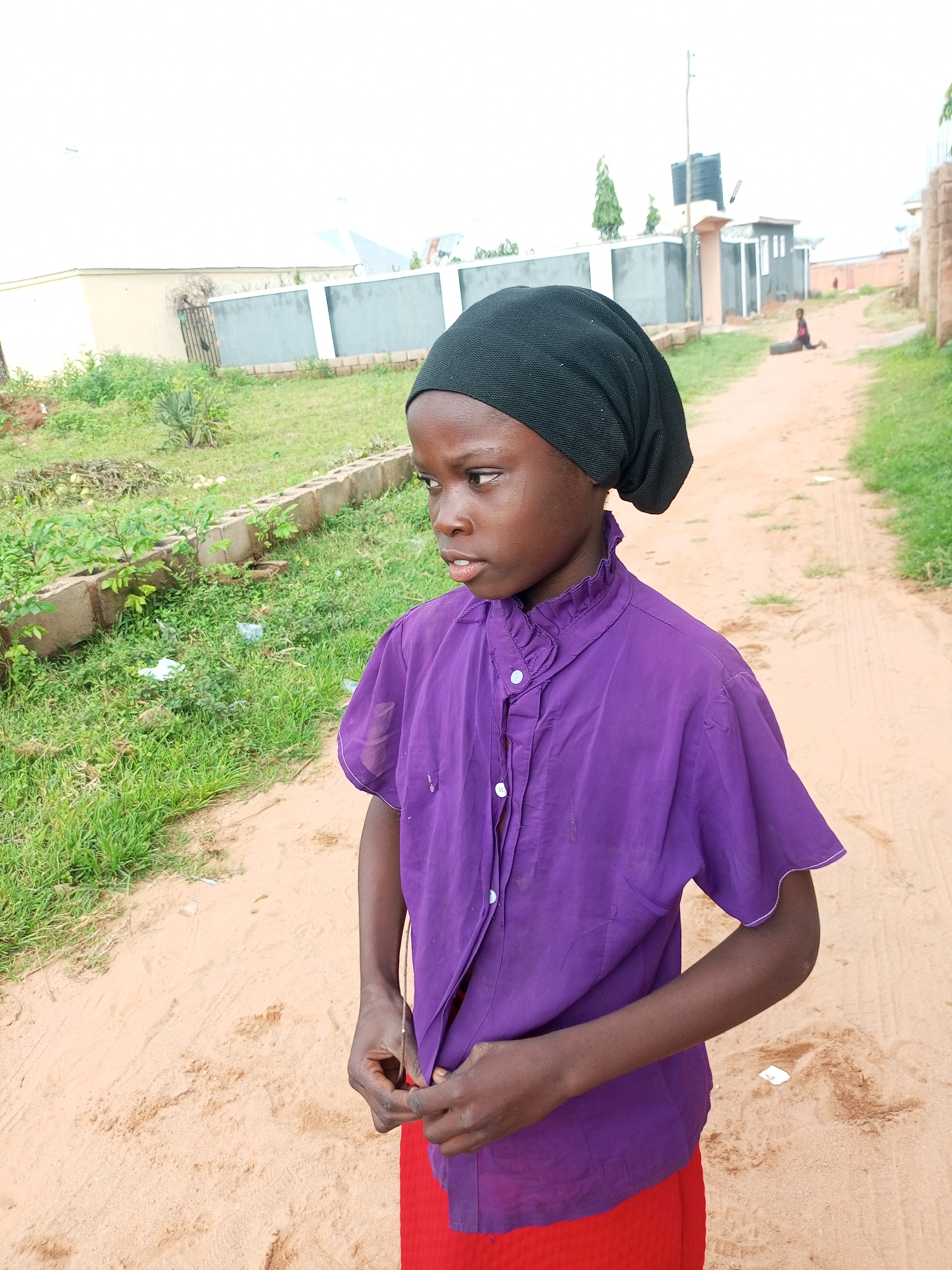 In Tempure, Nigeria, a young person stands in the middle of a sandy path, facing right with their body angled slightly away from the camera. They are wearing a purple button-up shirt with ruffled collar and white trim on the sleeves, and a red skirt. A black head covering is worn on their head. Their hands are near the bottom of their shirt. The path is bordered by sparse green grass and a low concrete barrier on the left, and more grass on the right. In the background, buildings with grey and white facades and a water tank are visible under an overcast sky. Another person is seated on the ground in the distance.