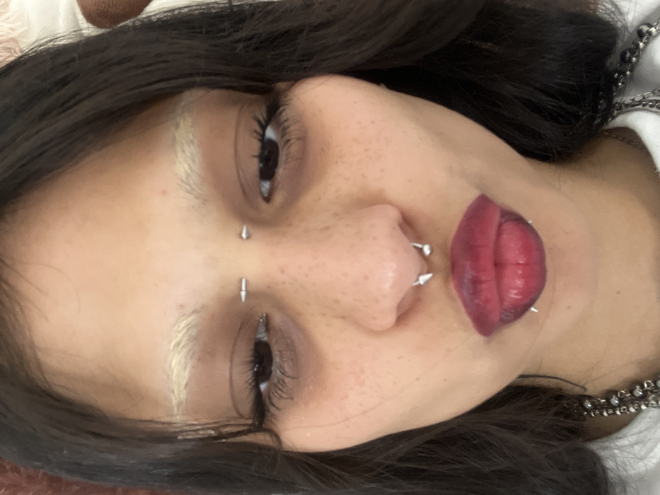 A person's face is displayed close-up, slightly rotated. They have dark hair and light eyebrows. The person has multiple facial piercings including one between their eyes, one under an eye, a ring through their nose and one through each side of their lower lip. The lipstick is dark red. The person has fair skin with freckles. A chain is visible around the neck.