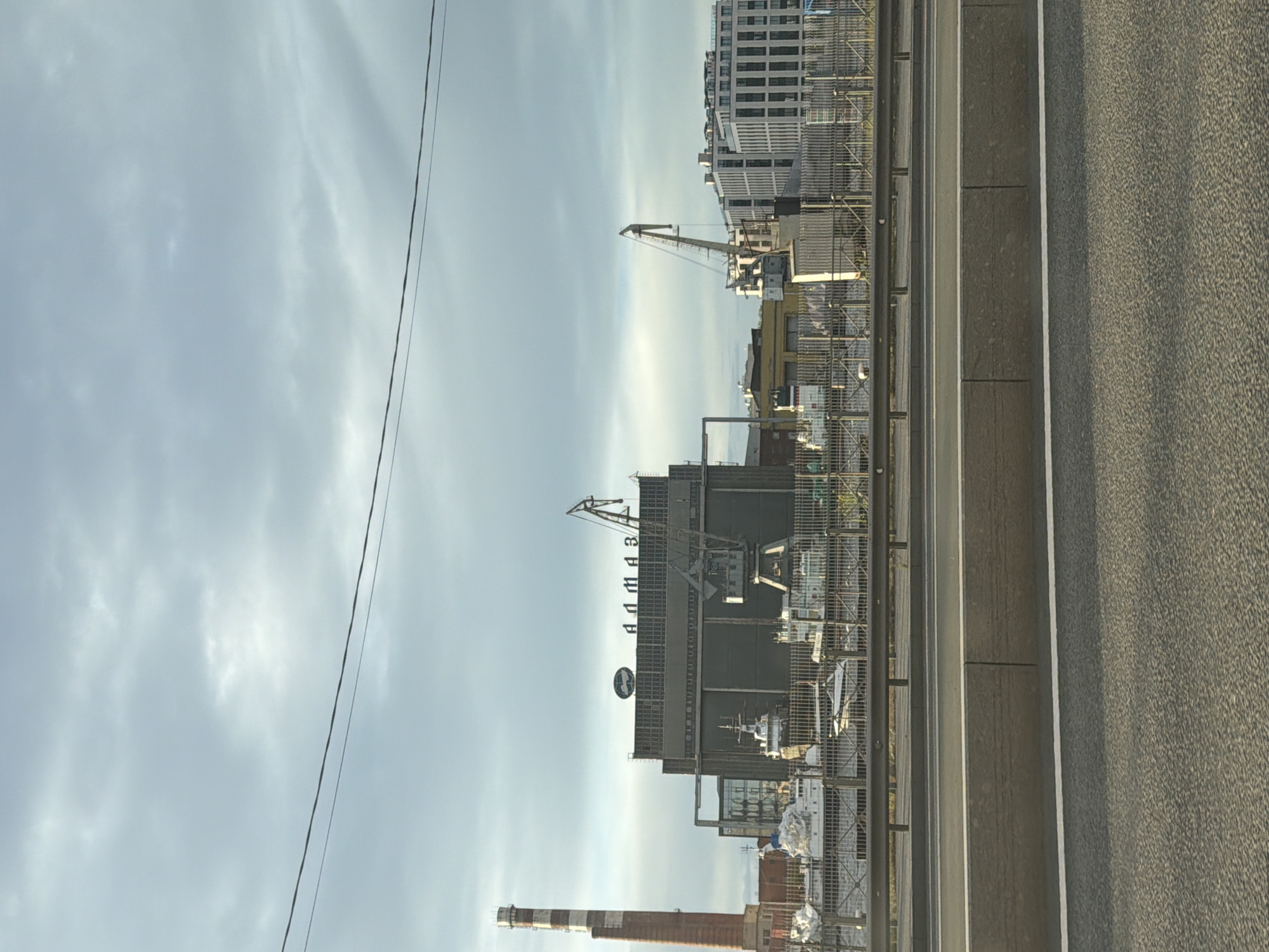 This is a photograph of a port with a cloudy sky and a road in the foreground. The image is oriented vertically, suggesting it might have been taken from a moving vehicle or held at an angle.
The subjects include several industrial objects: cranes, buildings, a tall smokestack, and possibly ships docked in the port. One prominent crane is visible in the middle ground, its arm extended. Behind it, a large building with "ANMAS" written in large letters across its facade is visible. Below this, a circular logo or sign is affixed to the building. Further to the right, another, taller building complex with many windows is visible.
The scene appears to be a working port. The presence of cranes and industrial buildings suggests an area dedicated to cargo handling or shipbuilding. The road in the foreground, marked with white lines, indicates it might be a road or highway adjacent to the port.
The time of day appears to be late afternoon or early evening, given the soft lighting and the color of the sky, which is a pale blue with significant cloud cover. The weather seems overcast but not actively raining. There are no people visible in the image.
The prominent text visible is "ANMAS" on a building. The overall impression is one of industrial activity in a coastal or riverside setting under a somewhat muted sky.
