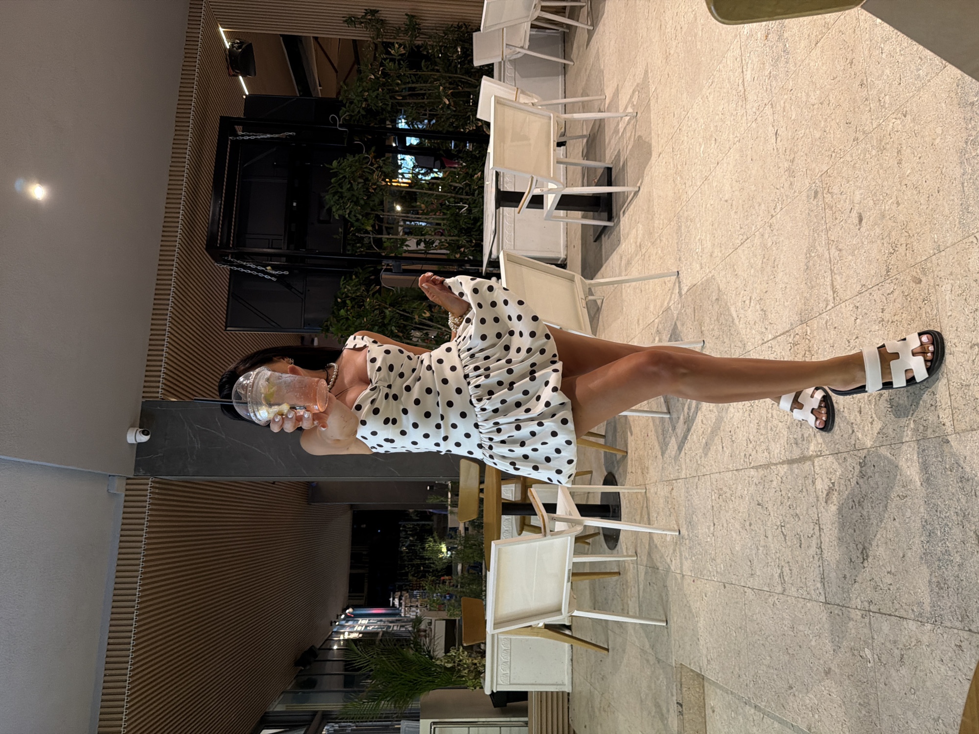 A woman stands in an outdoor restaurant area in Rafailovići, Montenegro, holding a drink. She is wearing a white strapless dress with black polka dots and white sandals with black soles. The restaurant has light-colored tiled flooring, and several white outdoor chairs and round tables are visible. In the background, there are potted plants and wooden architectural elements, including a slatted ceiling and vertical wooden panels. The lighting suggests it is evening or nighttime.