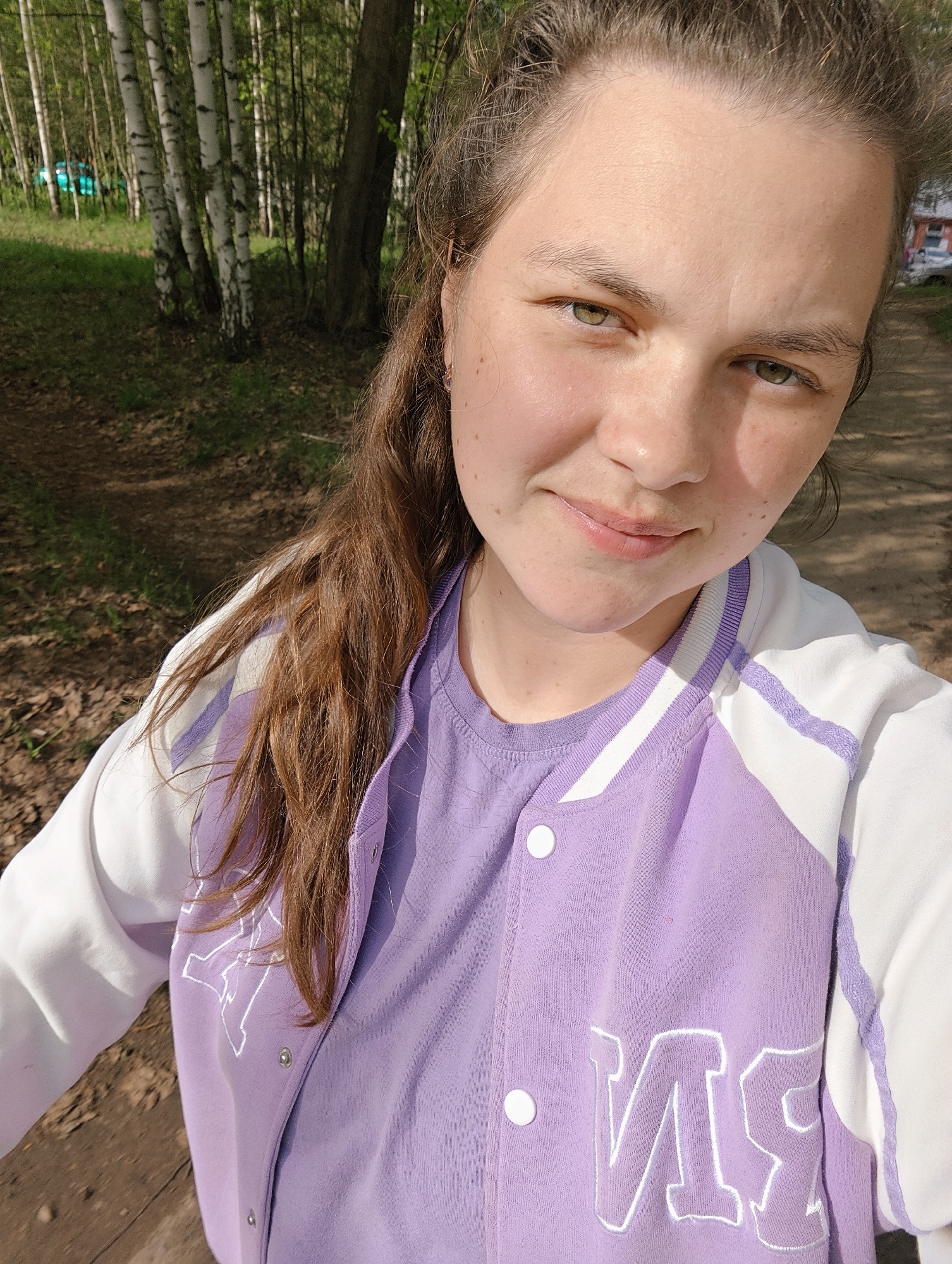A fair-skinned woman with brown hair is taking a selfie outdoors. She's wearing a lavender-colored t-shirt and a matching lavender and white varsity-style jacket. The background reveals a forest or park with birch trees. The location is likely in the city of Chaikovsky, Russia.