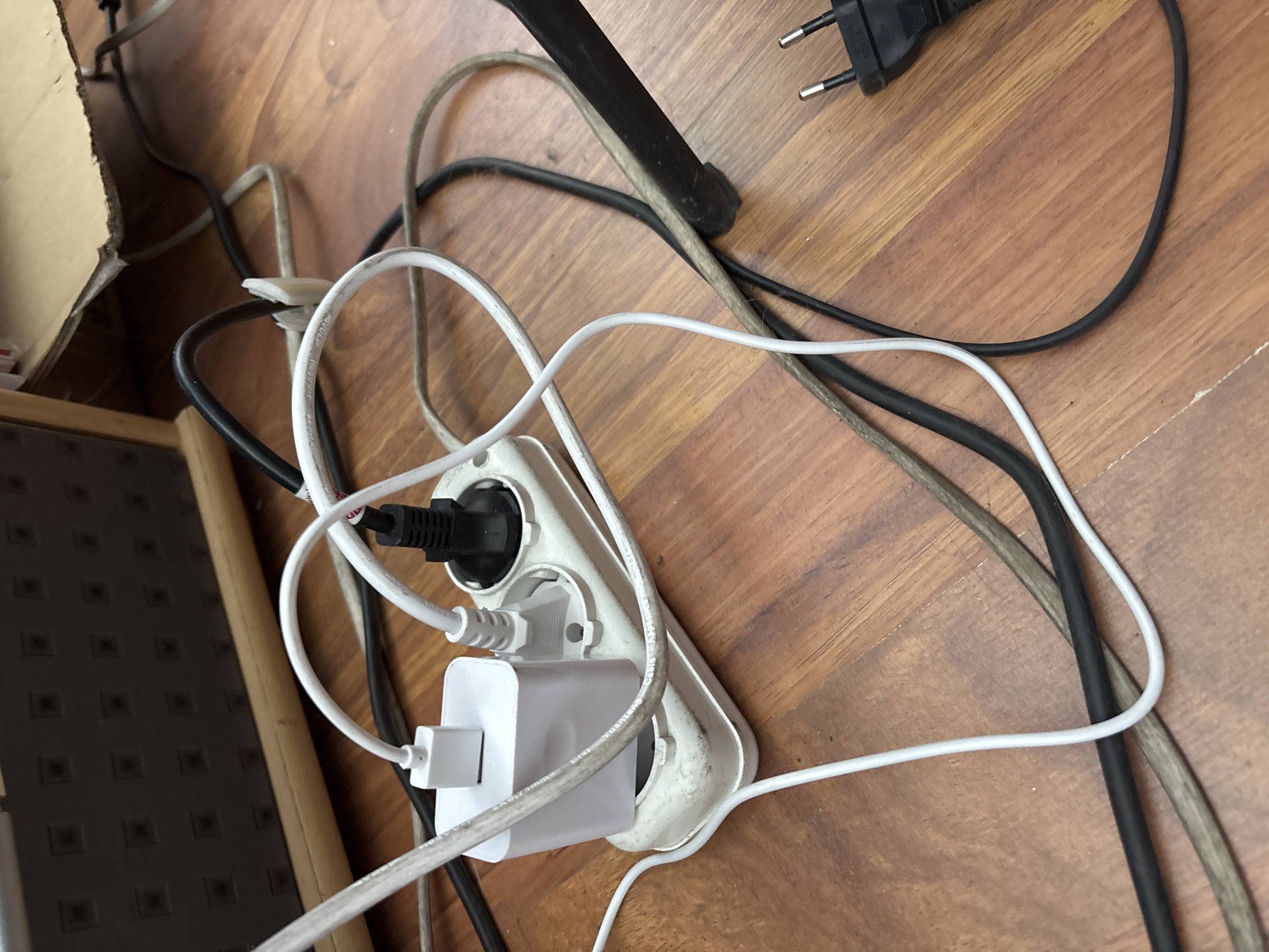 The image shows a close-up of a white power strip lying on a wooden floor. Several electrical cords are plugged into the power strip, including a white USB charger adapter and a black plug. The cords are tangled and spread across the floor. A portion of a dark object, possibly a piece of furniture or equipment, is visible on the left side of the image, and a cardboard box is visible in the background. The scene is indoors, and there is no indication of the specific location within Zaporizhzhia, Ukraine. The lighting appears to be artificial and somewhat dim.