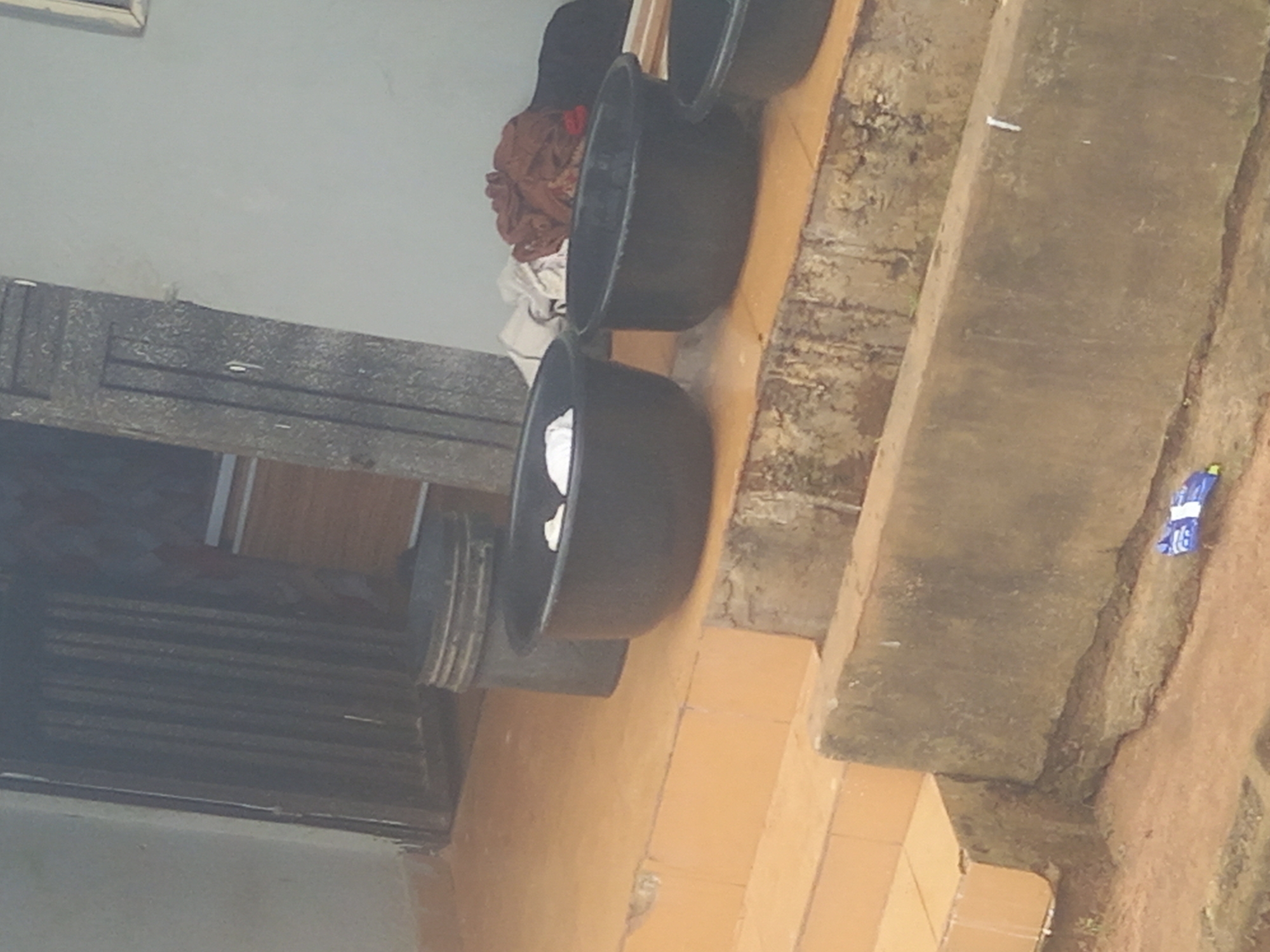 The image shows an outdoor scene with several black plastic basins placed on an orange tiled surface, which appears to be a set of steps or a patio. In the background, there is a dark gray, ribbed structure that could be a door or gate. Next to it is a large, dark gray cylindrical object, possibly a bucket or bin. A pile of clothing, including brown and dark fabrics, is visible behind the basins. The overall setting suggests a residential or utilitarian outdoor area. The ground in the foreground is a reddish-brown dirt surface, and a small blue object, possibly a piece of plastic packaging, is visible on a concrete ledge. The image is taken from a slightly elevated angle, looking down at the basins. There are no people visible in the image.
