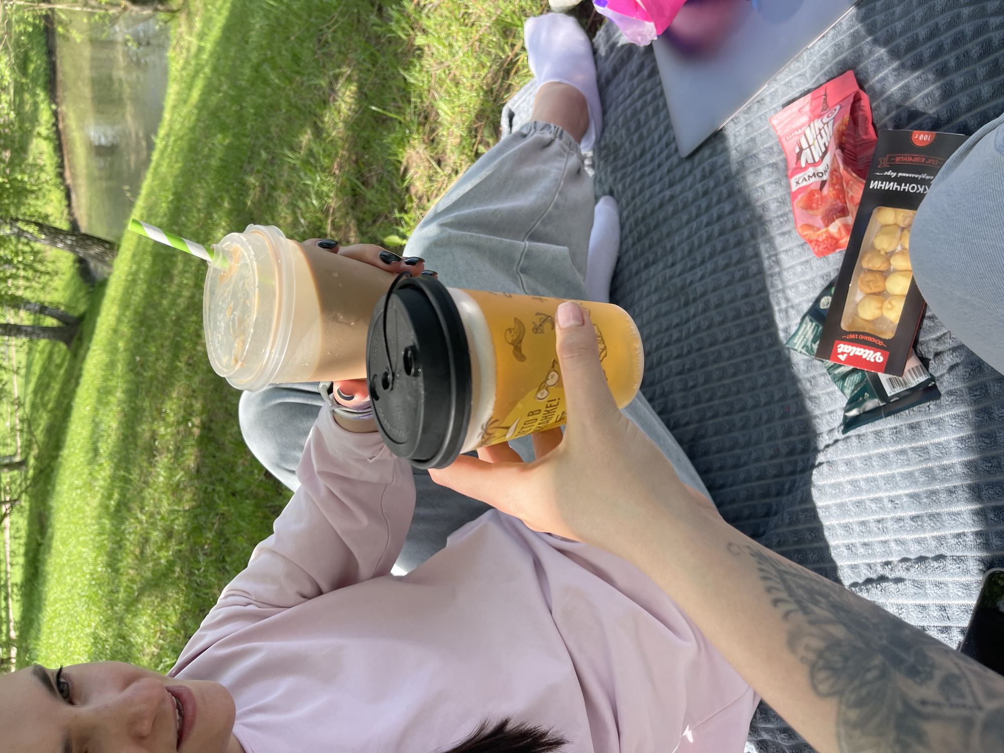 The image shows a picnic scene outdoors, with a woman holding two plastic cups with beverages. One cup contains a light brown liquid, possibly iced coffee, with a green and white striped straw. The other cup holds a yellow liquid. The woman is wearing a light pink long-sleeved shirt and grey sweatpants with white socks. She has a tattoo on her forearm.
A grey blanket is spread on the grass, and on it are a bag of "JAMON" flavored chips, a box of "Vitalat" cheese, and a smartphone. A grassy area with a body of water is visible in the background. The image suggests a casual, relaxed picnic outing. The location could be inferred as Moscow, Russia based on the Cyrillic text present on food packaging, the presence of a Vitalat product, and the packaging on the JAMON chips.