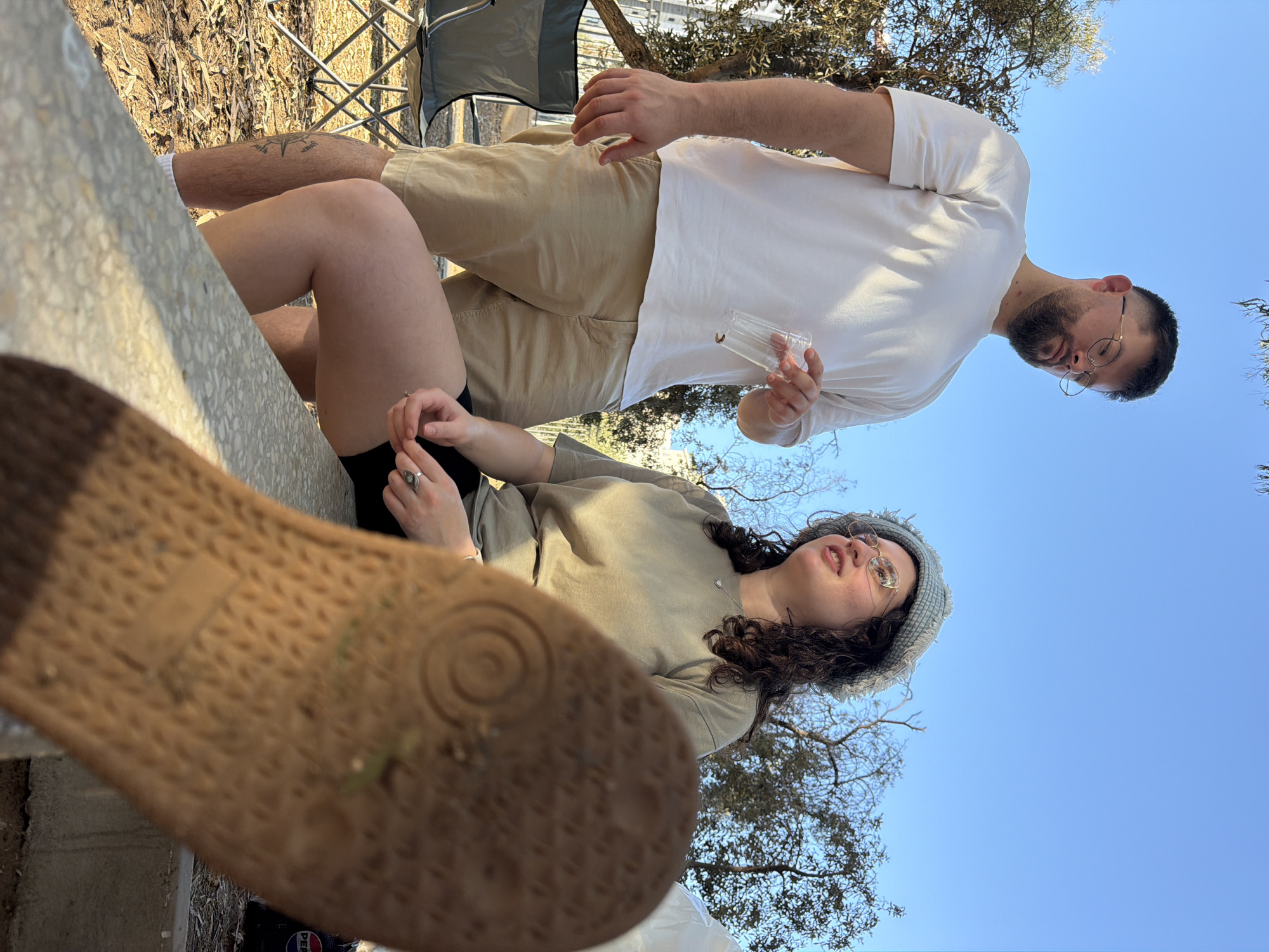 The image depicts a scene outdoors with two individuals under a clear blue sky. The individual on the left is a man with short dark hair and a beard, wearing glasses, a white t-shirt, and tan shorts. He is holding a clear plastic cup and looking down. The individual on the right is a woman with curly dark hair and glasses, wearing a light blue textured hat and a tan t-shirt. She is looking upwards and to the side, with her hands clasped in her lap. She is wearing black shorts. The foreground is dominated by the sole of a brown shoe, which is out of focus. They are seated on a stone bench. Behind them, trees and some foliage are visible, with a glimpse of buildings in the background. To the left, a camping chair is partially visible. A black bag with a red and blue logo, possibly a Pepsi bag, is seen on the right. The setting appears to be a park or an outdoor recreational area. The location is identified as Rishon LeZion, Israel.