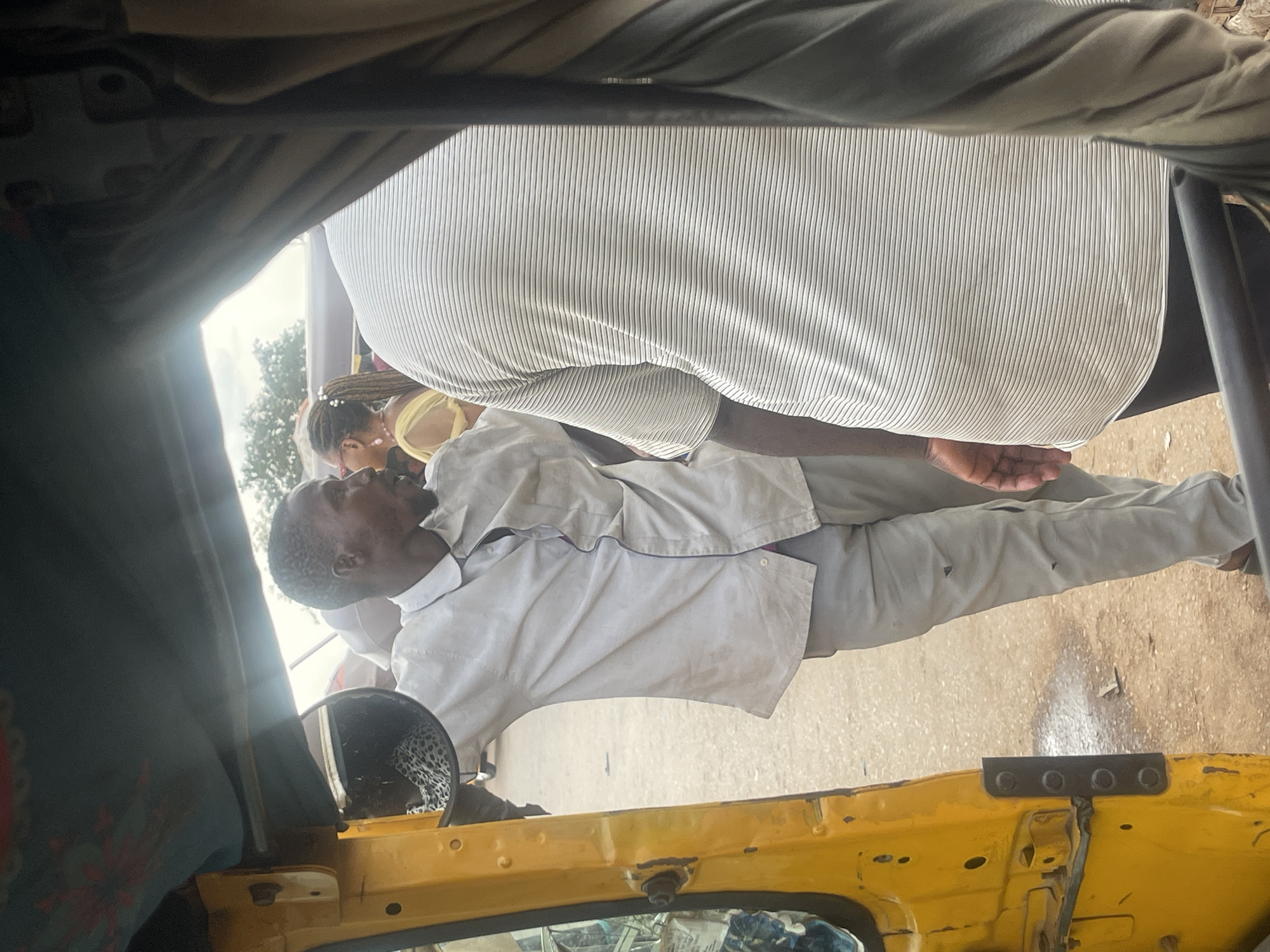 In Galadimawa Village, Nigeria, a man in a light-colored collared shirt and light-colored pants leans against the open door of a yellow auto-rickshaw. His left arm is resting on the door frame, and his right hand is visible near his chest. A second person, wearing a horizontally striped t-shirt, is positioned to the right, facing away from the camera. In the background, a woman with braided hair adorned with beads and wearing a yellow garment is visible, along with trees and overcast sky. The ground is paved with a mix of sand and dirt.