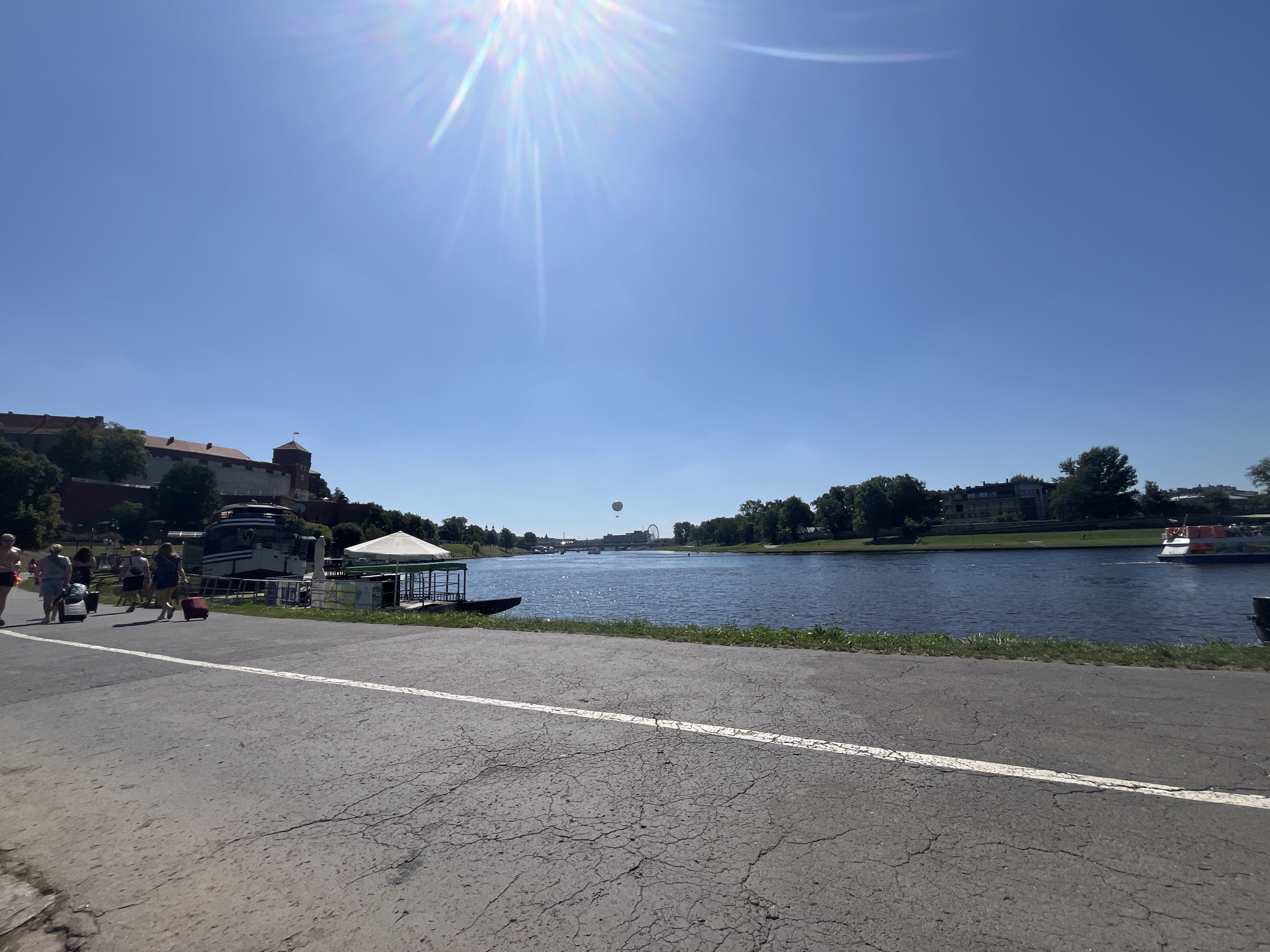 The image depicts a scenic view of the Vistula River in Krakow, Poland, on a bright, sunny day. On the left bank, a section of Wawel Castle is visible, characterized by its red-tiled roofs and a prominent tower. A pathway runs alongside the river, where several individuals are seen walking, some pulling suitcases, suggesting they are tourists. A large tour boat is docked near the pathway, alongside a covered seating area. The river itself is calm, reflecting the clear blue sky and the bright sun, which creates lens flare. Across the river, the opposite bank is lined with lush green trees and buildings. In the distance, a Ferris wheel and a hot air balloon are visible, adding to the lively atmosphere of the cityscape. A smaller passenger boat is cruising on the river. The foreground shows a cracked asphalt pathway with a white line marking its edge.