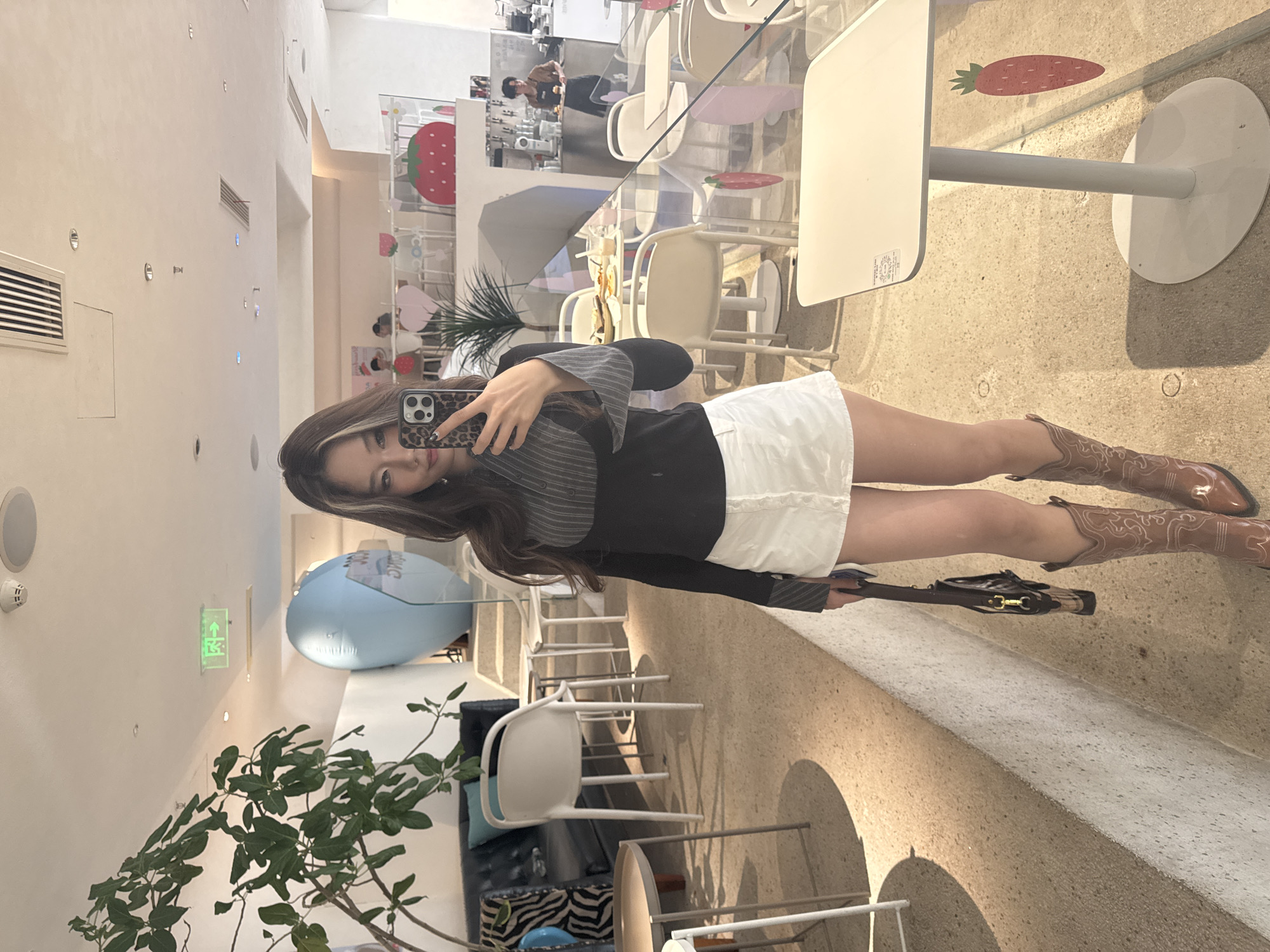 Here is a forensic description of the image:
The image captures a woman taking a selfie in an interior space, likely a café or restaurant, in 江北城街道, China. The woman is wearing a striped long-sleeved top, a white skirt, and brown cowboy boots. She is holding a brown handbag. Her phone has a leopard print case. In the background are tables with decorations, including a strawberry and a carrot image, and several chairs. The interior design is light and modern with white walls and some decorative elements such as a blue balloon and a potted plant. Other patrons can be seen in the background, suggesting a public commercial space.