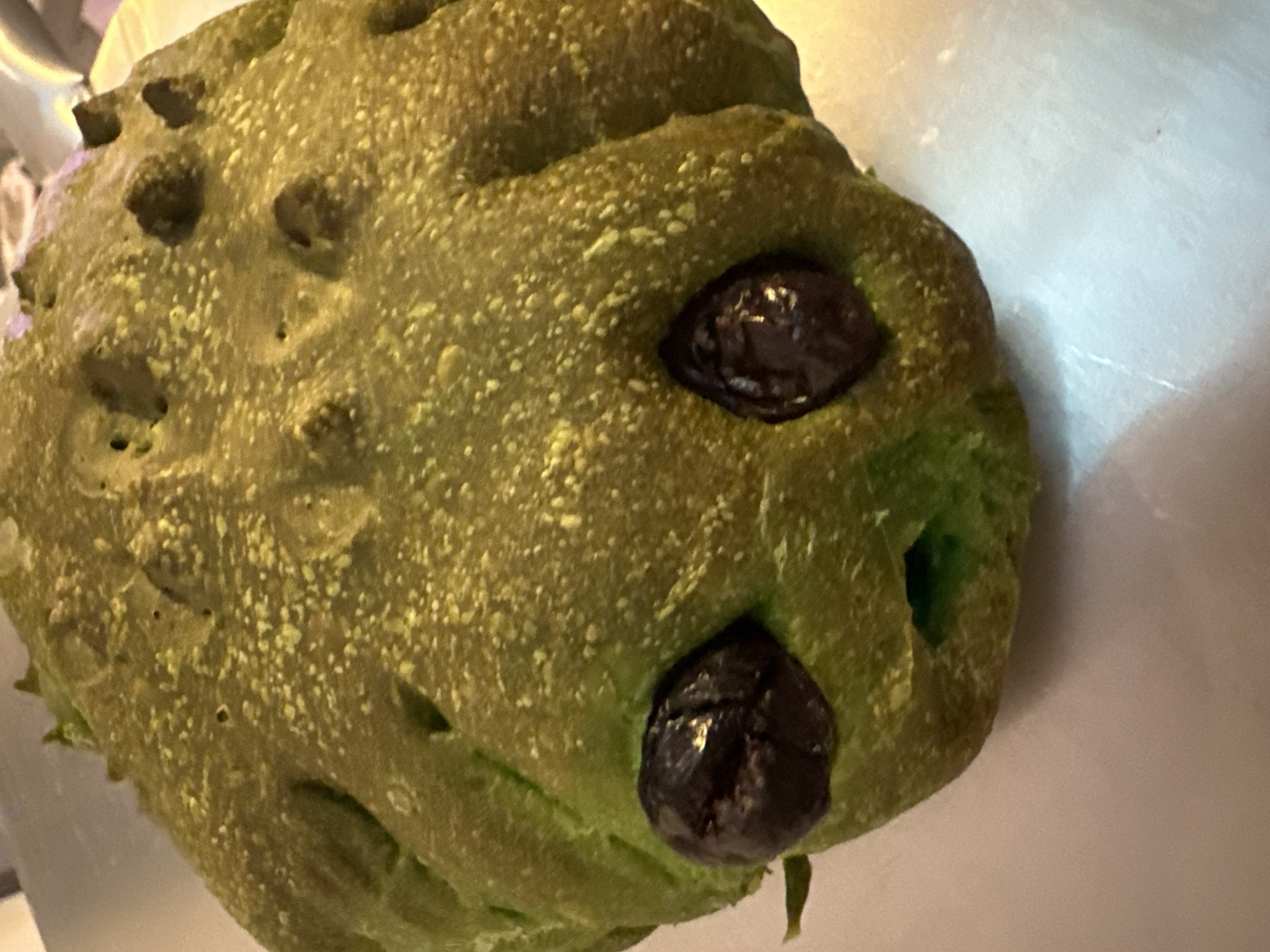The image shows a close-up of a green, olive-studded bread shaped like a creature, possibly an octopus or frog, on a light-colored, textured surface. The bread has two dark, round olive eyes and a small, open mouth, giving it a somewhat surprised or distressed expression. The surface it rests on appears to be a metallic or plastic tray. The lighting casts a slight shadow, highlighting the texture and bumps on the bread. There is no indication of the city of Moortown, Ireland, or any other specific location.