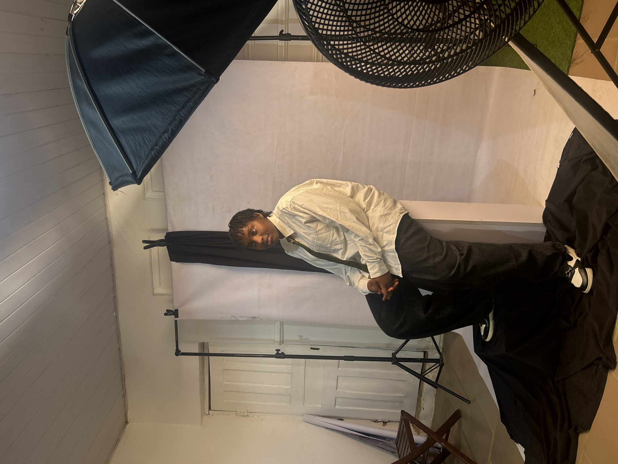 The image shows a person with short dark hair, wearing a white long-sleeved shirt and dark pants, leaning against a white backdrop. The person is looking at the camera with their hands clasped. The setting appears to be a photography studio, evident from the presence of lighting equipment like a softbox and a fan. The background includes a white wall and a door. A black cloth is draped on the right side of the image. The location, Jos, Nigeria, is not determinable from the image.