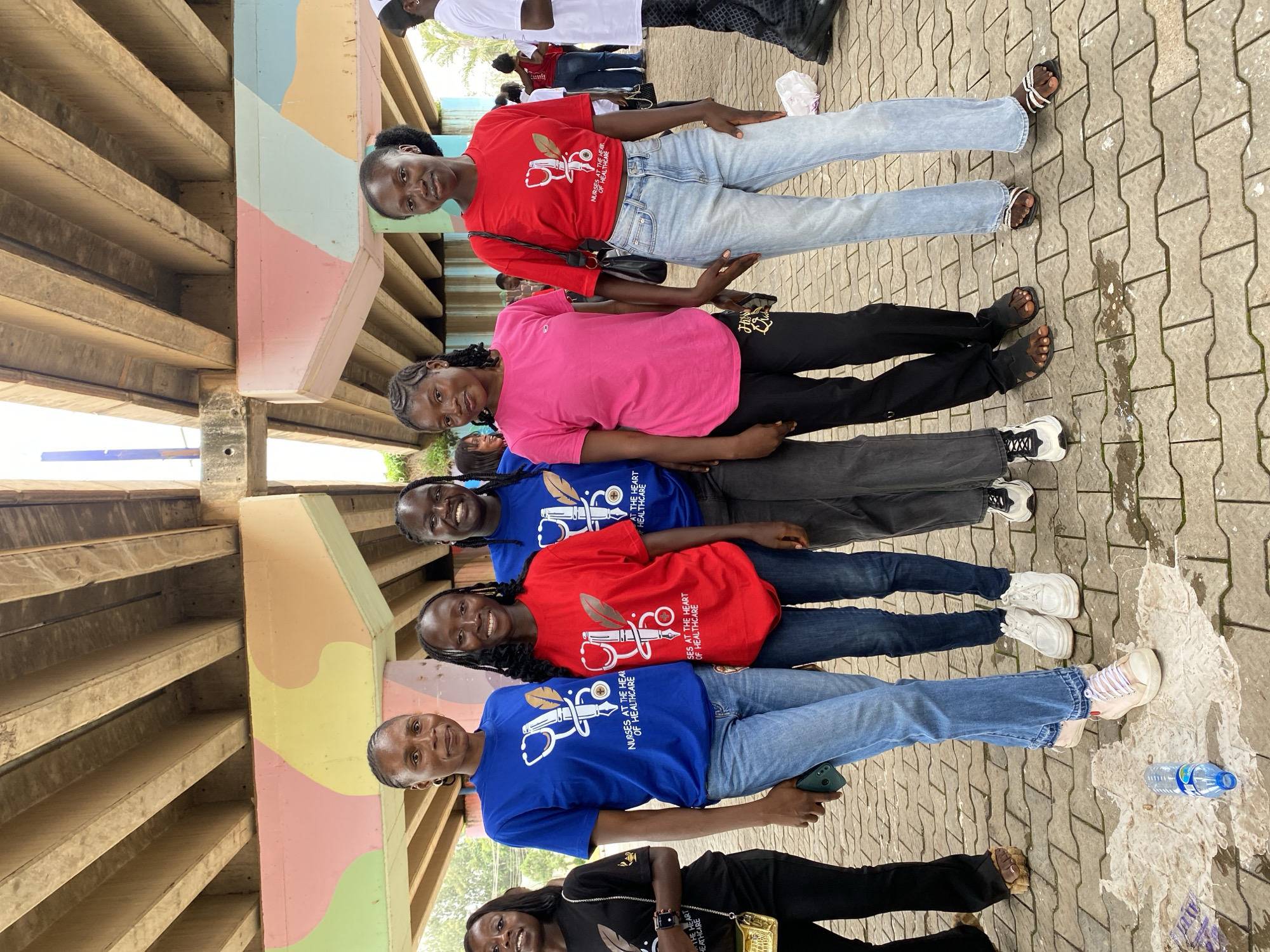 A group of young women, likely students or professionals in healthcare, are posing for a photo under a concrete overpass or bridge. The overpass features colorful murals on its support columns in shades of pink, yellow, and light blue. The women are standing on a paved ground that appears wet in places.
Several of the women are wearing matching blue t-shirts with white text and graphics that include a stethoscope and quill pen. One woman in the front is wearing a black t-shirt with similar graphics and text. Other women are wearing red and pink t-shirts, and most are dressed in jeans. Their footwear includes sandals and sneakers.
The overall mood appears positive, with several women smiling at the camera. The lighting suggests it is daytime, and the wet ground could indicate recent rain or a spilled drink. The presence of a water bottle suggests they might be on a break or attending an outdoor event. The scene is informal, with the women gathered together for a casual group photograph. The text on the blue shirts clearly reads "NURSES AT THE HEART OF HEALTHCARE," identifying them as nursing professionals or students.