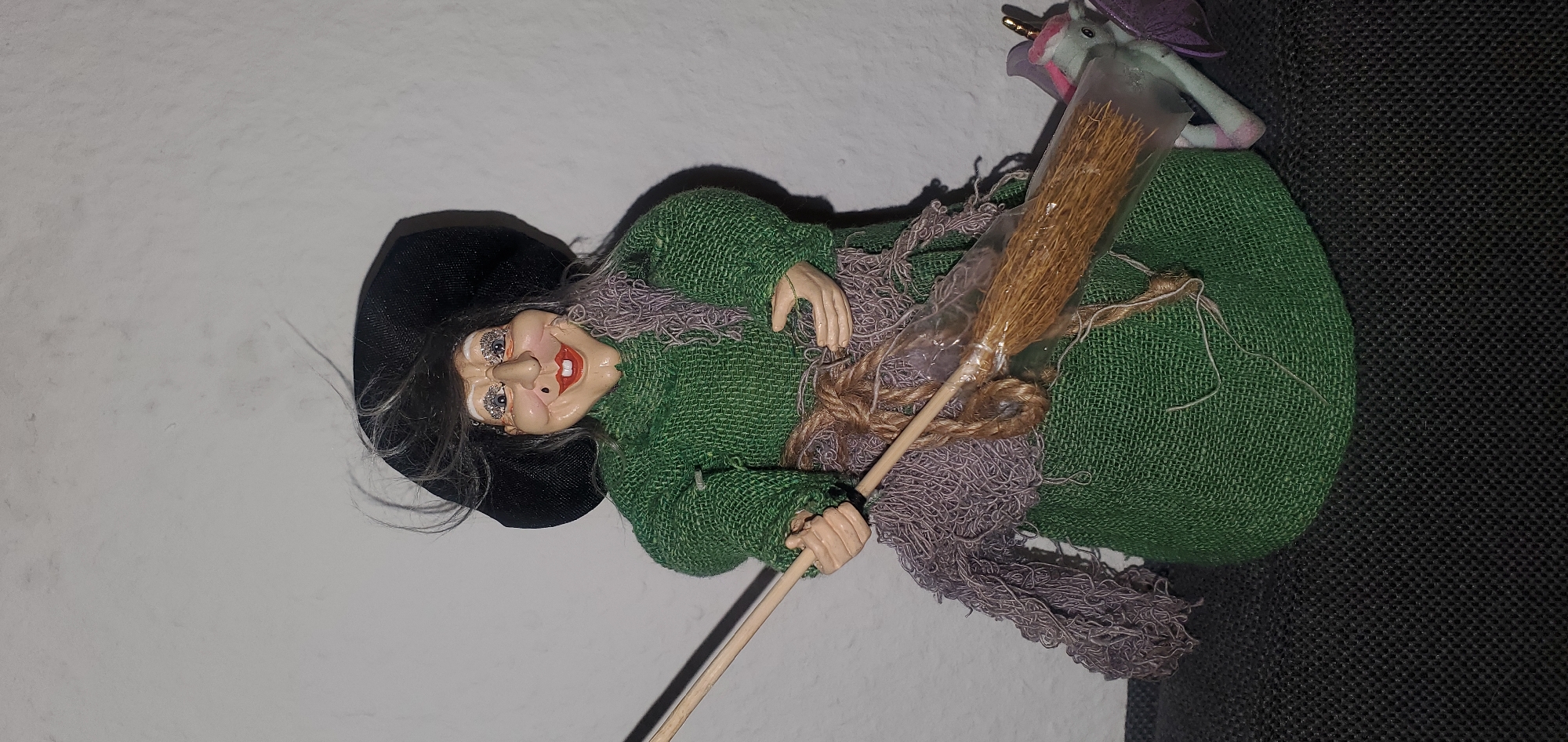 A doll resembling a witch stands in front of a plain white wall, its body draped in a green fabric garment. The witch wears a black hat and a purple scarf. A small broom is held in its hands, attached to a larger object that appears to be a unicorn figure. The background is a white wall, and the foreground is a dark, textured surface. There is no indication of the doll's location in Sasbach, Germany.