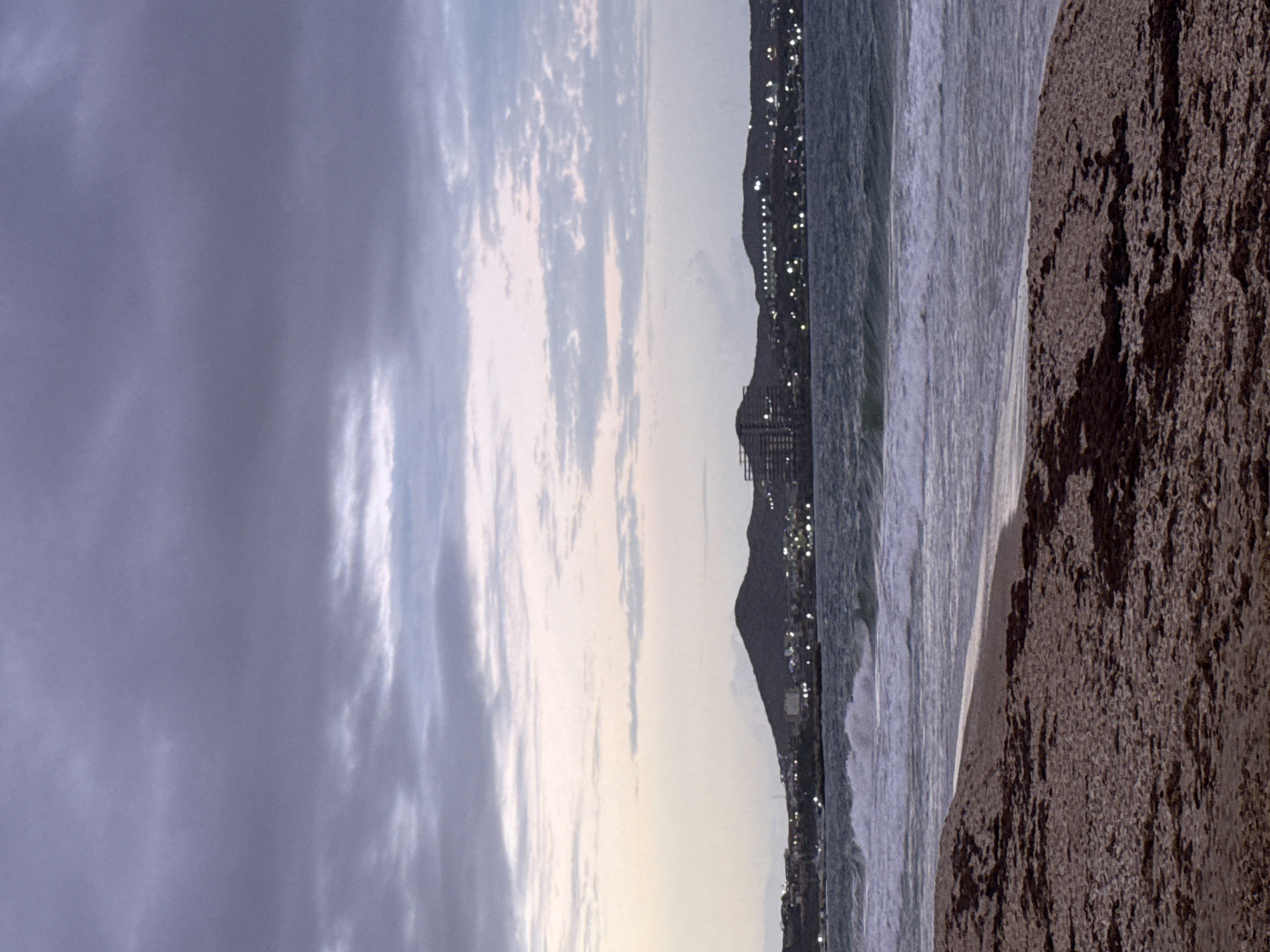 The image depicts a serene coastal landscape captured at dusk or night, likely in Porlamar, Venezuela. The sky occupies a significant portion of the upper frame, characterized by dark, purplish-grey clouds that transition to lighter, almost iridescent pale blue tones on the right, suggesting the last vestiges of daylight or moonlight.
Below the sky, a distant, undulating coastline of hills or mountains stretches across the middle of the image. This landmass is illuminated by countless small, bright artificial lights, twinkling like stars against the dark terrain, indicating a sprawling city or town along the shore.
Adjacent to the illuminated coastline, the ocean features prominently. Dark, cool-toned waters show visible waves with white foamy crests as they break, reflecting the low light. To the right, a wide expanse of sandy beach fills the lower part of the frame. The sand appears dark brown and coarse-grained, with discernible textures and small shadows.
The scene conveys a tranquil, nocturnal ambiance, highlighting the contrast between the natural elements of sky, sea, and sand, and the distant glow of human settlement. No people or discernible text are visible in the image.