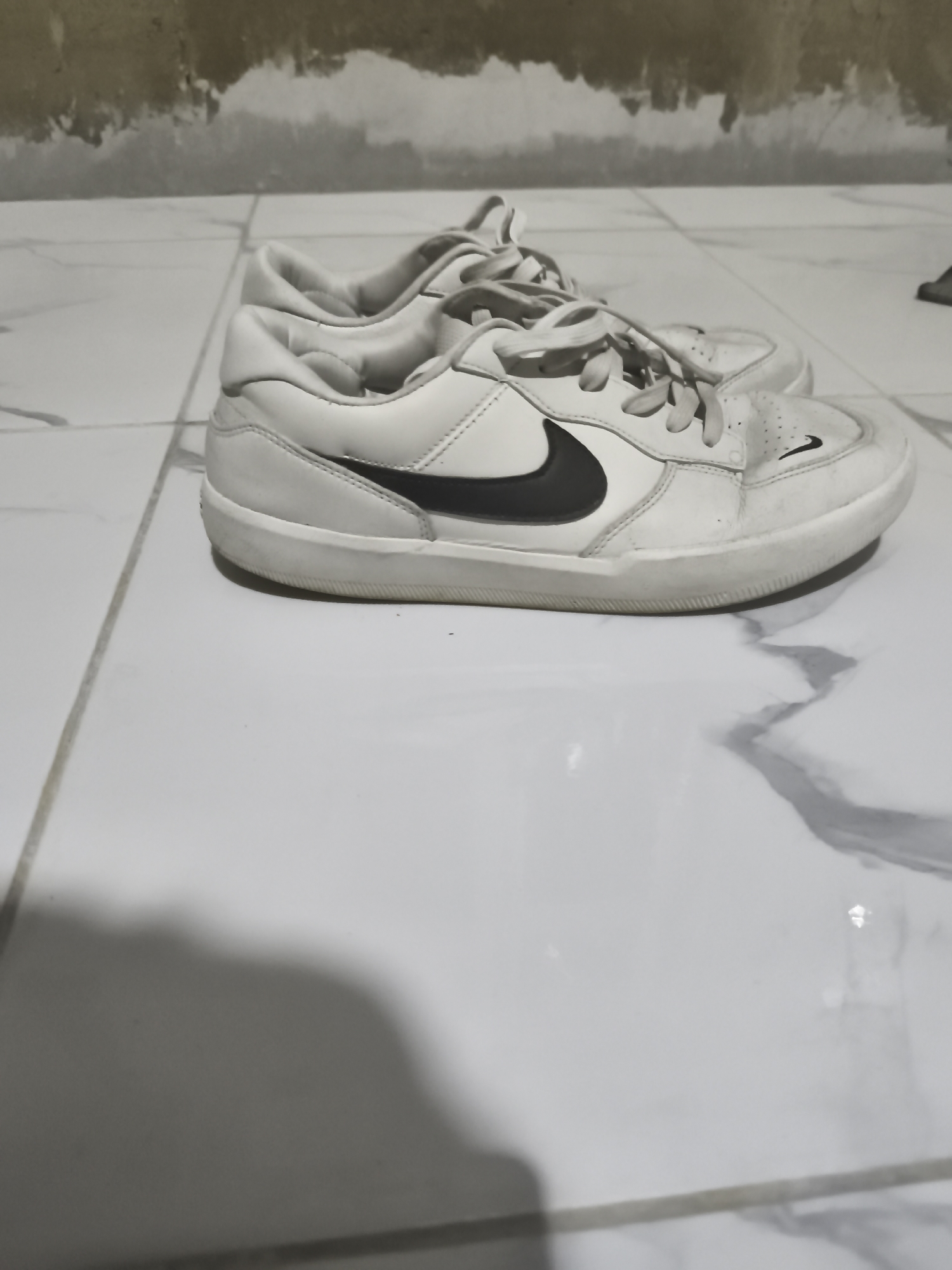 A pair of white Nike sneakers with black swoosh logos is placed on a tiled floor. The sneakers appear slightly worn. The background shows a textured, possibly concrete wall with some peeling paint or discoloration. The floor tiles are large and white with grey veining, characteristic of marble or a similar stone. The lighting suggests an indoor setting, likely a room with artificial light, casting a shadow of the shoes on the floor. The overall scene is a simple still life of footwear.