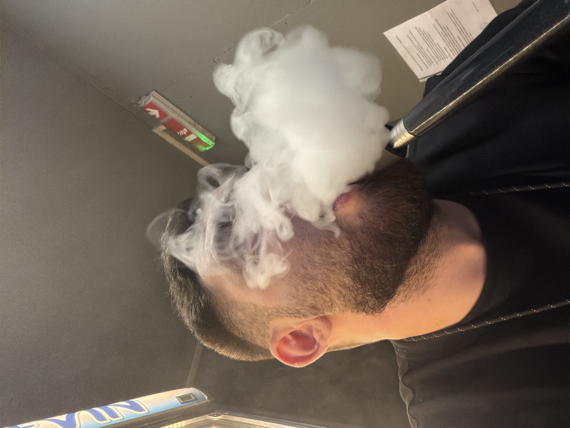 A low-angle shot depicts a man with a beard and close-cropped dark hair exhaling a large plume of white smoke, possibly from a vaping device or hookah. The environment is dimly lit. An exit sign with a red arrow and green indicator light is visible on the ceiling. A metal or plastic object with blue and white lettering is partially visible in the lower-left corner. The man is wearing a black shirt. The image was most likely taken inside, based on the ceiling and lighting. I cannot infer a location in Varna, Bulgaria.