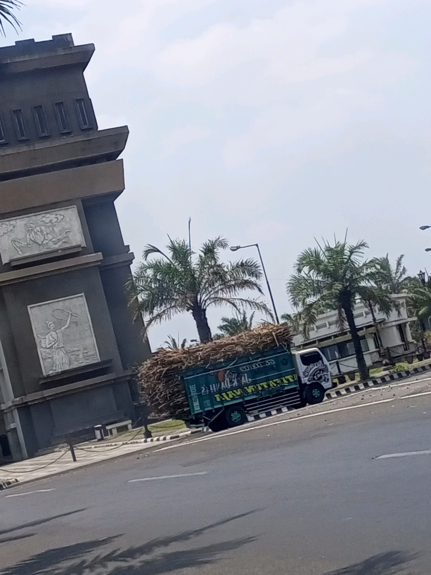 A truck loaded with agricultural products, possibly sugarcane, drives on a street in Gurah, Indonesia. The truck is teal and white, with various decals on its side. It is surrounded by palm trees, and a large concrete structure with carvings is visible on the left. The sky is overcast.
