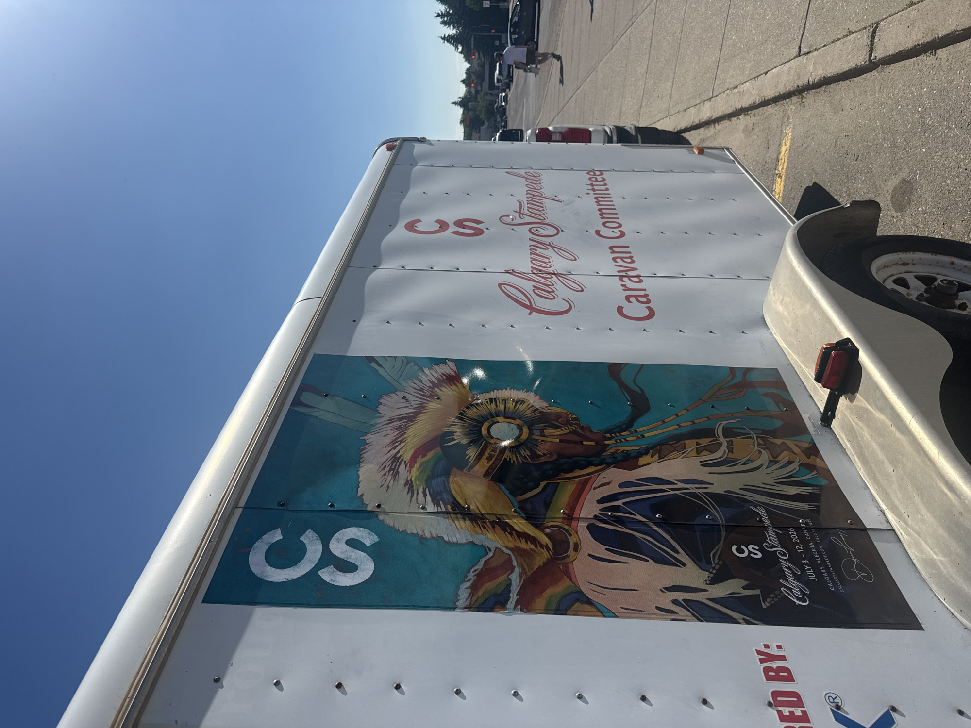 A white trailer with the Calgary Stampede logo and artwork of a person in headdress is parked on the side of a city street. The trailer is white and has multiple rivets across its surface. On the side of the trailer, there are two instances of "CS" in red lettering, one larger and one smaller. Below the larger "CS" are the words "Calgary Stampede" in a cursive red font, followed by "Caravan Committee" in a smaller red font. To the left of this, a large graphic depicts a person's head and shoulders adorned with a colorful feathered headdress and braids. The graphic has a teal background. Below this graphic, the text "RED BY:" and a blue logo are partially visible. The trailer has a fender over a wheel. In the background, there is a sidewalk, a street with cars, traffic lights, and trees. A man in a white shirt and shorts is walking on the sidewalk. The sky is clear and blue.