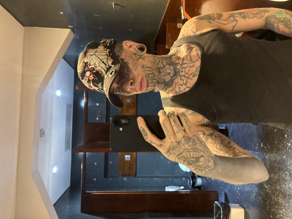 An individual with extensive facial and body tattoos, appearing to be male, is captured in a mirror selfie within an indoor setting. The subject is wearing a black ribbed tank top, an ornate black and multicolored baseball cap featuring graphic designs and metallic eyelets, and possesses large earlobe gauges. Facial tattoos include script on the jawline and patterns resembling stitches or lines around the eyes. Neck and arm tattoos are elaborate, featuring floral, geometric, and script designs. The subject holds a black smartphone in the right hand, angled to capture their reflection.
The background depicts a dimly lit, dark blue or teal room with numerous square indentations or marks on the walls. Dark wooden partitions or stall doors are visible to the left and right of the subject. A white-lined waste bin sits on the dark floor to the far left, which appears to have some light-colored debris. The ceiling is light-colored with two visible recessed spotlights and a rectangular ventilation grate. The environment suggests a public restroom or changing area.
The specific location within the city of Buenos Aires, Argentina, cannot be determined from the visual information provided in the image.