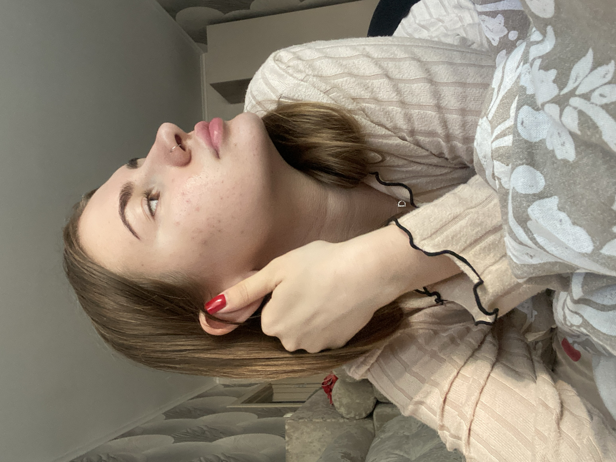 This is a close-up, vertically oriented, indoor photograph of a young person with light skin, light brown hair, and light brown eyes. The person is lying down, with their head tilted upwards and to the left, obscuring much of their body. They are wearing a light-colored, ribbed long-sleeved shirt with black trim at the cuffs. A grey blanket or comforter with a white floral pattern is visible on the right side of the frame, partially covering the person.
The person's face is the primary focus. They have a nose ring, and their lips are slightly parted. The skin on their face shows some texture and visible pores. Their right hand is raised, with bright red nail polish on the fingernails, and the index finger and thumb are touching their ear and hair, respectively. The lighting suggests it might be daytime, possibly in a bedroom or living area, as indicated by the soft, diffused light and what appears to be furniture in the background. The overall mood is relaxed and intimate. There is no visible text in the image.