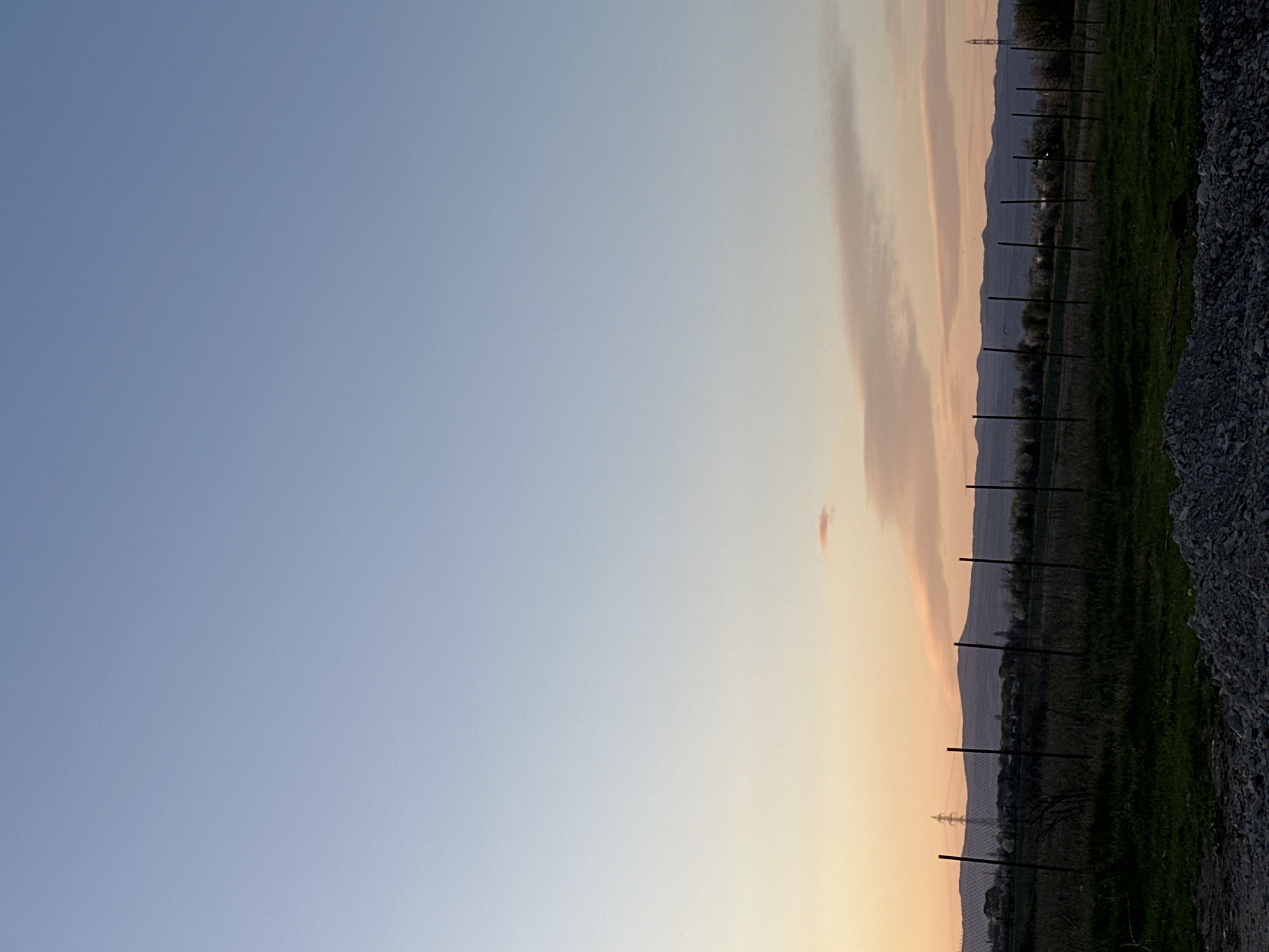 The image depicts a landscape scene at sunset. The sky transitions from light blue to orange-pink hues. There is a strip of land with a green grassy field and a line of fence posts running along it. In the distance, there are hills. Power lines are visible in the background, near the hills. The foreground to the right contains gravel or small rocks. Location cannot be definitively determined as Sofia, Bulgaria from the image.