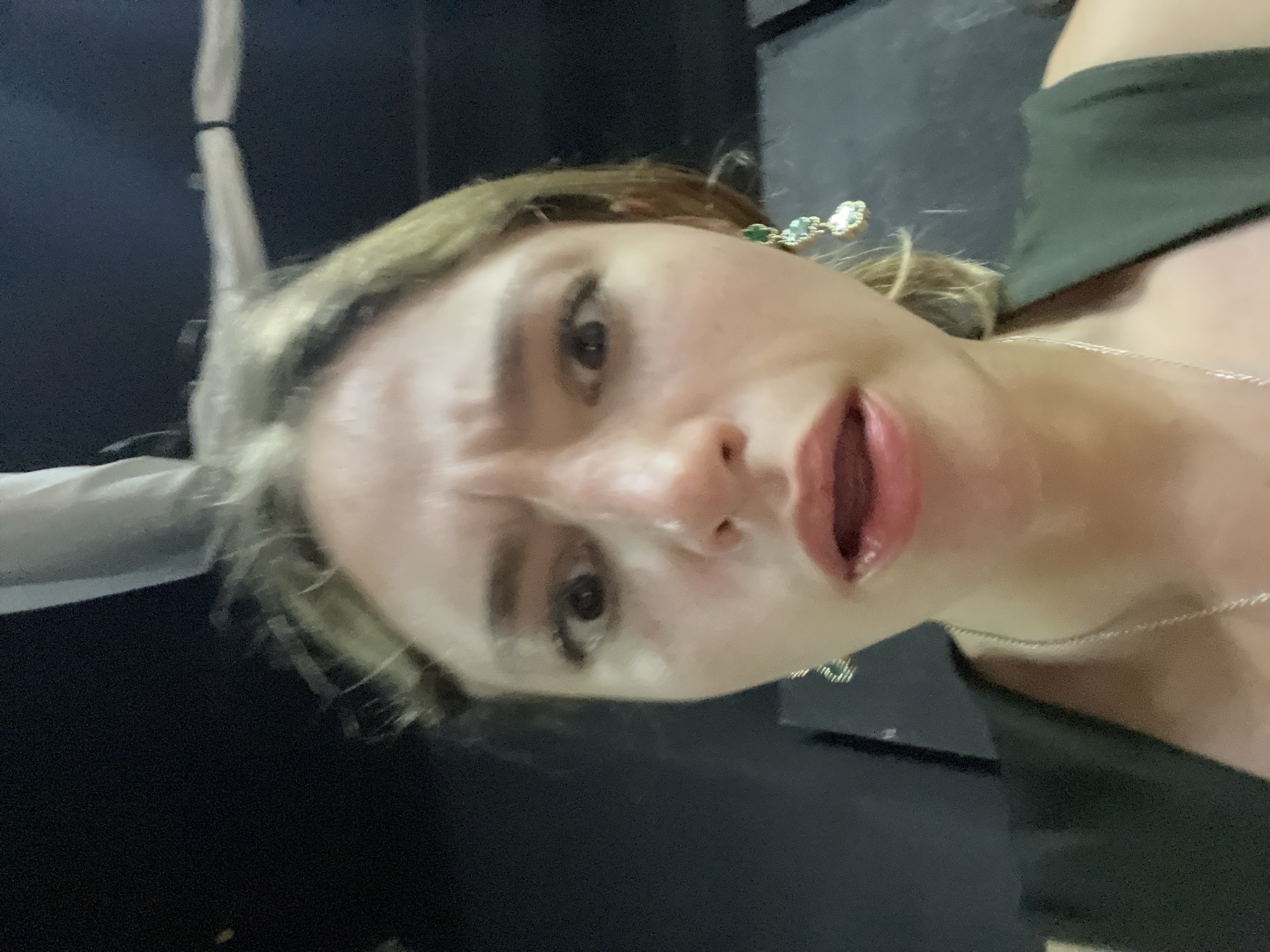 This is a close-up, first-person perspective photograph of a young woman with light brown hair, wearing a dark green sleeveless top and a silver necklace. She is looking directly at the camera with wide eyes and a slightly open mouth, her eyebrows furrowed as if in surprise or concern. She has on dangling earrings with green and light blue stones. The background is dark and out of focus, with hints of what appears to be industrial piping or rigging overhead. The lighting is dim, with some light reflecting off her forehead and cheeks. There is no indication in the image itself of the location being San Bernardino Tlaxcalancingo, Mexico; this information is provided externally.