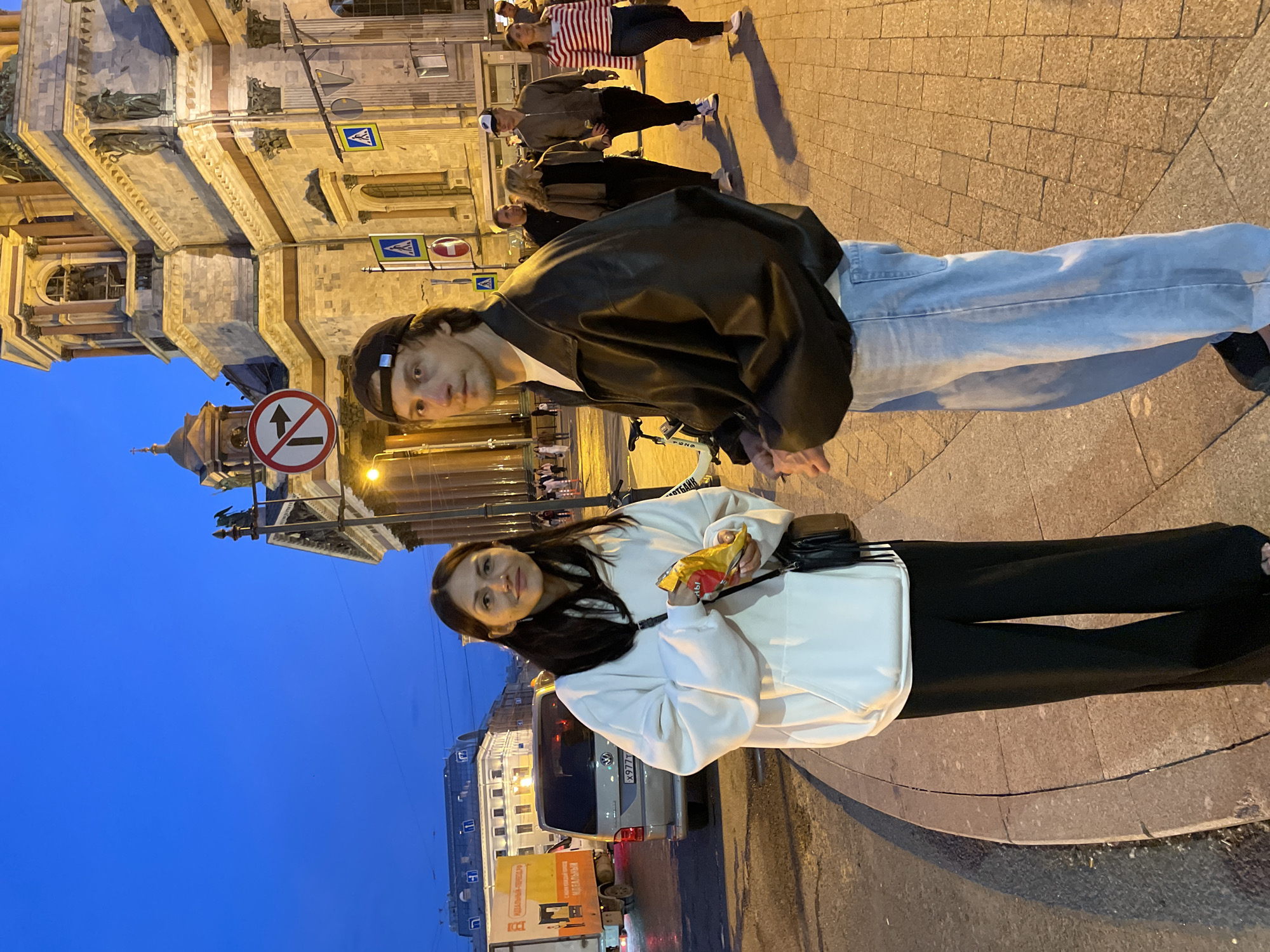 In an urban setting at dusk, a young Caucasian couple stands on a cobblestone sidewalk, illuminated by streetlights and the warm glow of nearby buildings. The woman, on the left, wears a white hooded sweatshirt and black wide-leg pants, holding a bag of chips in her right hand. She has long, dark hair and a friendly expression. The man, on the right, sports a black leather jacket, a black baseball cap, and light blue jeans. He looks directly at the camera with a neutral expression. Behind them, a large, ornate building with classical architecture dominates the background, its facade lit by artificial light. Several other pedestrians and a silver van are visible in the midground and background, suggesting a busy public space. A traffic sign with a red circle and arrow indicates a no-left-turn restriction. The sky above is a deep blue, transitioning from day to night.