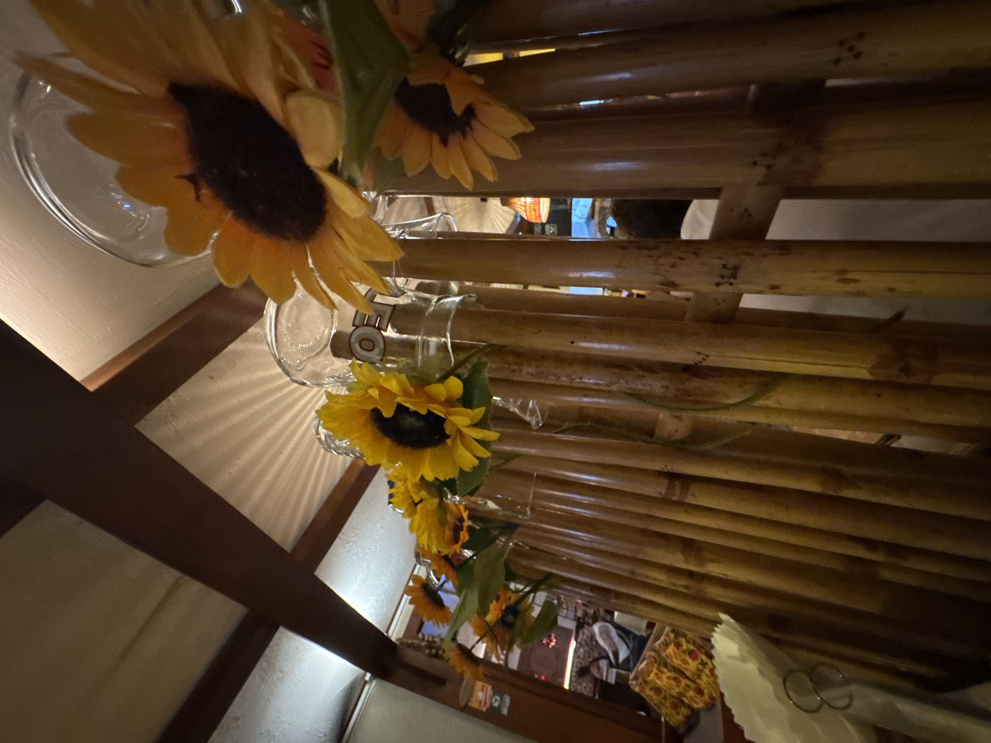 The image shows a close-up, slightly tilted view of a table decorated with sunflowers. The sunflowers, with their bright yellow petals and dark centers, are arranged in glass vases. The table itself appears to be made of bamboo, with horizontal slats that create a textured surface.
In the background, glimpses of a room are visible. There are wooden beams, possibly part of a ceiling or wall structure, and light filtering in from an unseen source, creating a patterned illumination on a light-colored surface. Further in the background, through the bamboo slats, there are hints of other elements, including what looks like fabric with a patterned design, and possibly other people or figures, though they are not clearly discernible. The overall impression is one of an indoor setting, perhaps a restaurant or a home, decorated for a casual or festive occasion. The lighting suggests it could be daytime, or an indoor setting with warm artificial lighting. No text is visible in the image.