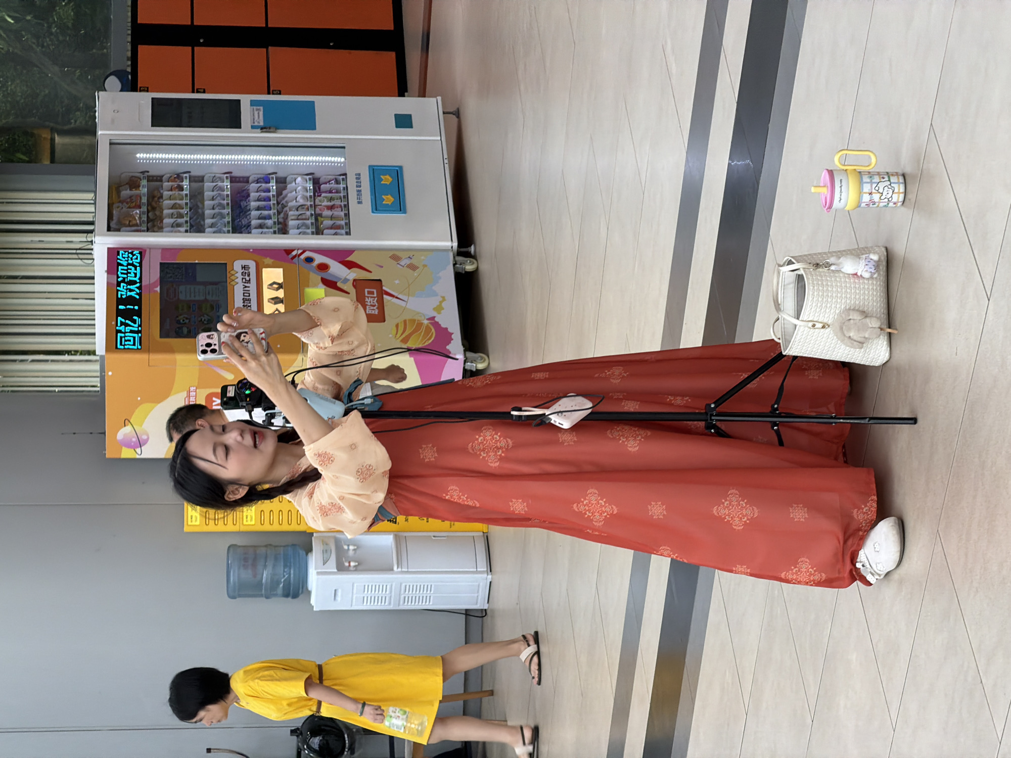 In an indoor public space with tiled flooring, likely a commercial area or transit hub in Jiangbeicheng Subdistrict, China, a young woman in a flowing red and gold traditional Chinese dress is recording herself with a smartphone mounted on a tripod. She holds the phone in her right hand and appears to be speaking to the camera. Her hair is styled in braids. In the background, to the left, another woman wearing a bright yellow dress walks away from the camera, holding a bottle of water. Behind the woman recording, a large vending machine with an illuminated display offering snacks and drinks is visible, along with a water cooler to its left. To the right of the vending machine, orange lockers are stacked. In the foreground, next to the tripod, a woven white handbag with two plush charms hangs, and a colorful child's sippy cup sits on the floor. The overall scene suggests a casual, public setting where the woman is engaged in content creation.