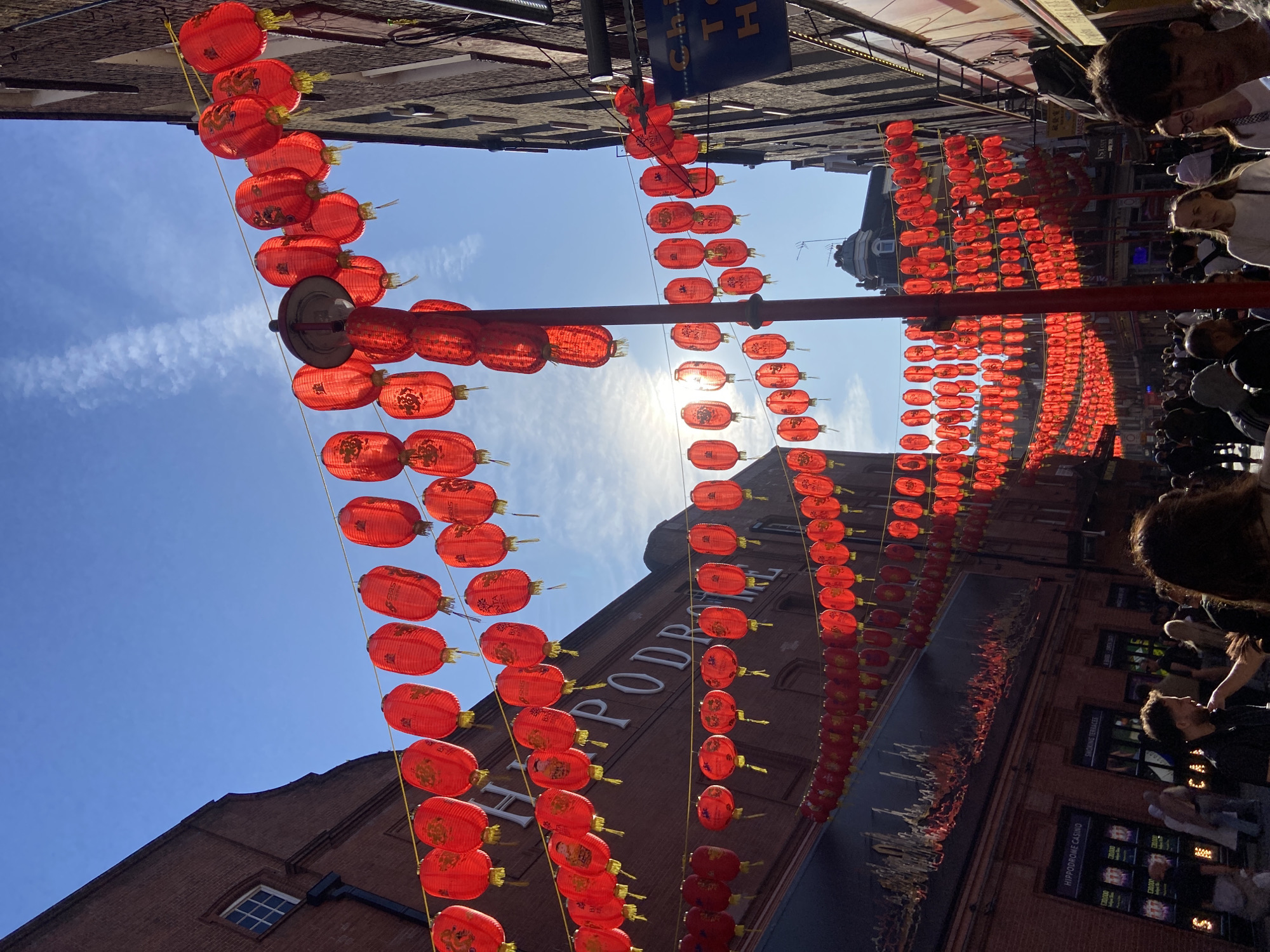 The image shows a street in London's Chinatown, adorned with rows of red Chinese lanterns strung overhead. Sunlight breaks through the lanterns, creating a bright, airy atmosphere. People are walking along the street, engaging in conversations and observing the surroundings. The buildings lining the street display signs and advertisements, including one for the Hippodrome Casino. The sky is clear with a few scattered clouds.