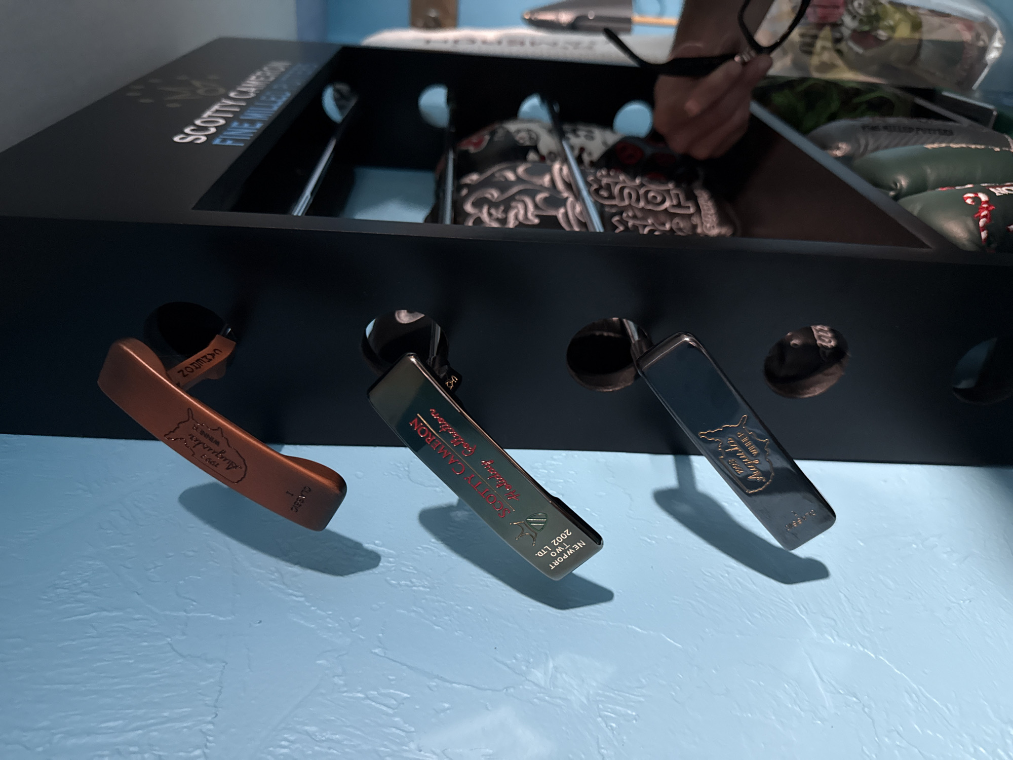This image shows a display of Scotty Cameron putter headcovers in what appears to be a retail or private collection setting. The headcovers are inserted through holes in a black display case, with a line of putter covers visible inside the display. The headcovers on display are a mix of designs, including what appear to be limited edition or tour-specific models. The colors vary, from a copper tone to black and gray. The image captures a sense of collecting and a display of golf equipment. The exact location in North Vancouver, Canada, cannot be determined from the image alone.