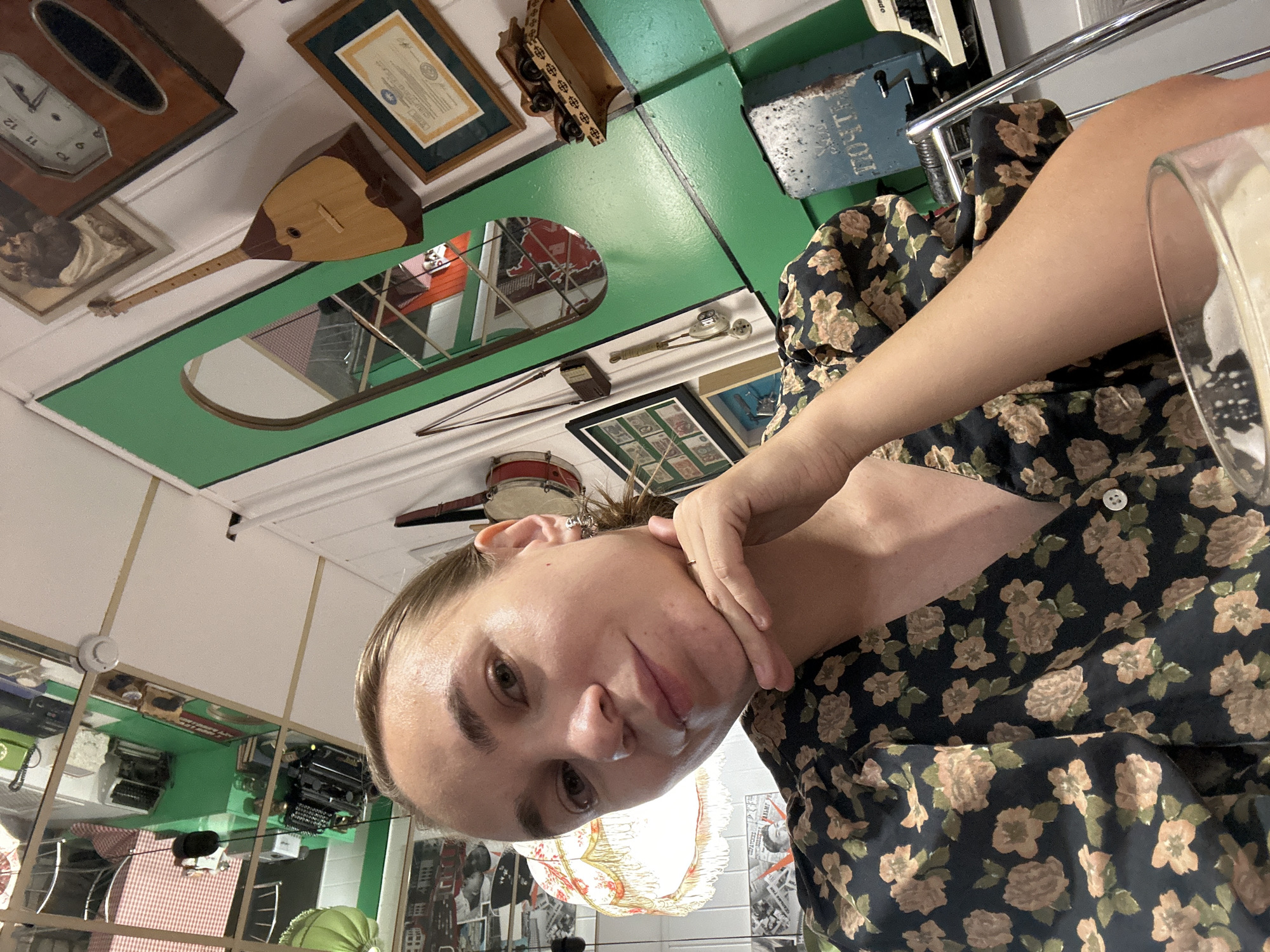 A woman with blonde hair pulled back is taking a selfie in what appears to be a quirky, eclectic cafe or shop. She is wearing a dark floral print dress. Her right hand is resting on her chin. The background is filled with various objects, including a mirror reflecting the ceiling which has what looks like a checkered pattern and numerous items, a wooden stringed instrument, a clock, framed pictures and documents, a red drum, and a vintage-style blue postal box. The overall impression is a cluttered but charming environment. This location is consistent with the interior of a cafe in Elista, Russia, known for its unique cultural influences and artistic expressions.