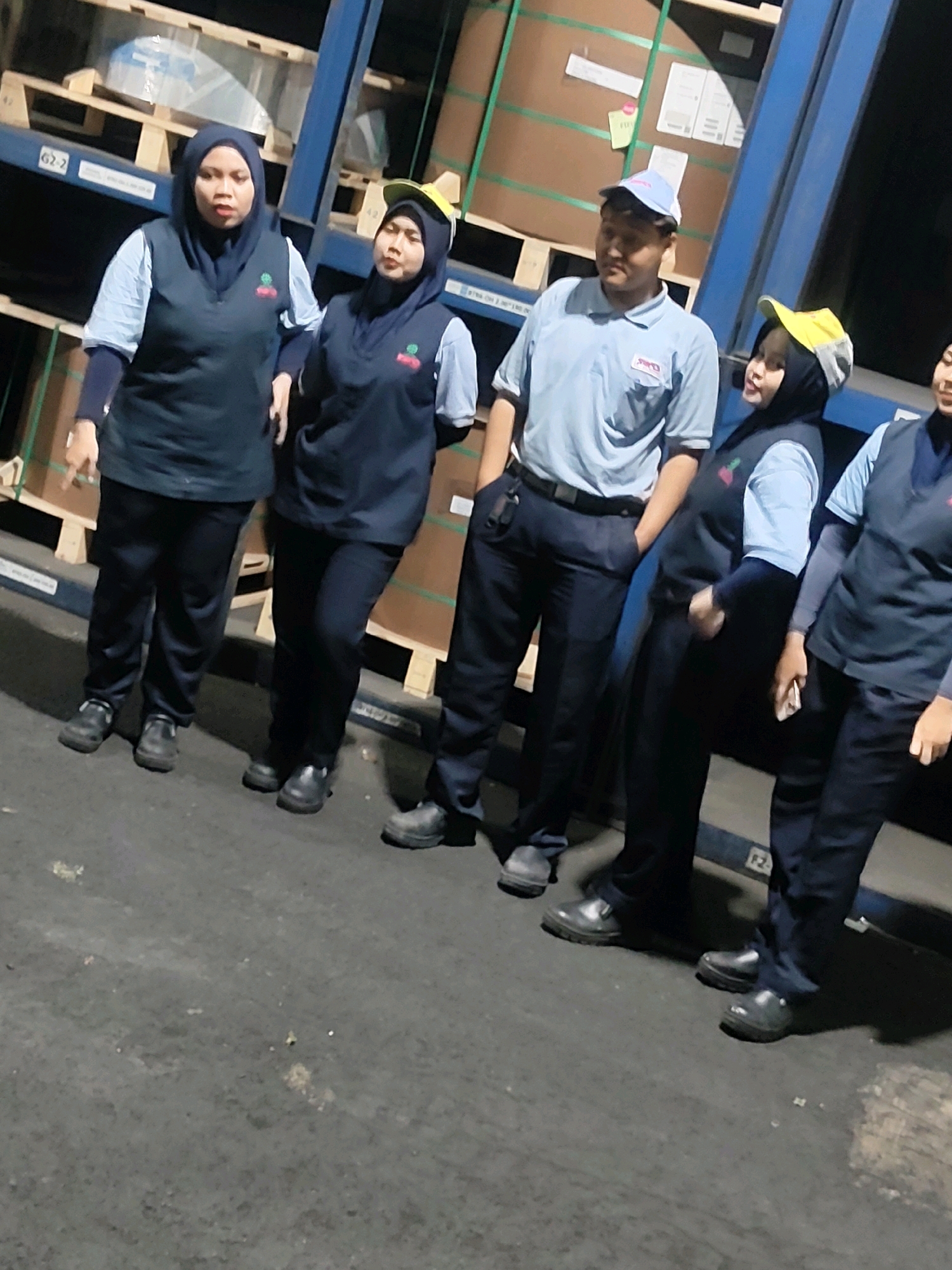 The image shows a group of five individuals, four women and one man, posing for a photo in a warehouse setting. The individuals are wearing dark blue uniforms with light blue long-sleeved shirts underneath. The women are also wearing hijabs, two in navy blue and two in yellow and navy blue. The man is wearing a light blue polo shirt and a light blue cap. They are standing on a dark gray concrete floor. In the background, there are tall shelves filled with large rolls of paper or similar material. The overall scene suggests a workplace environment, likely a distribution or manufacturing facility. The lighting appears to be artificial and somewhat dim.