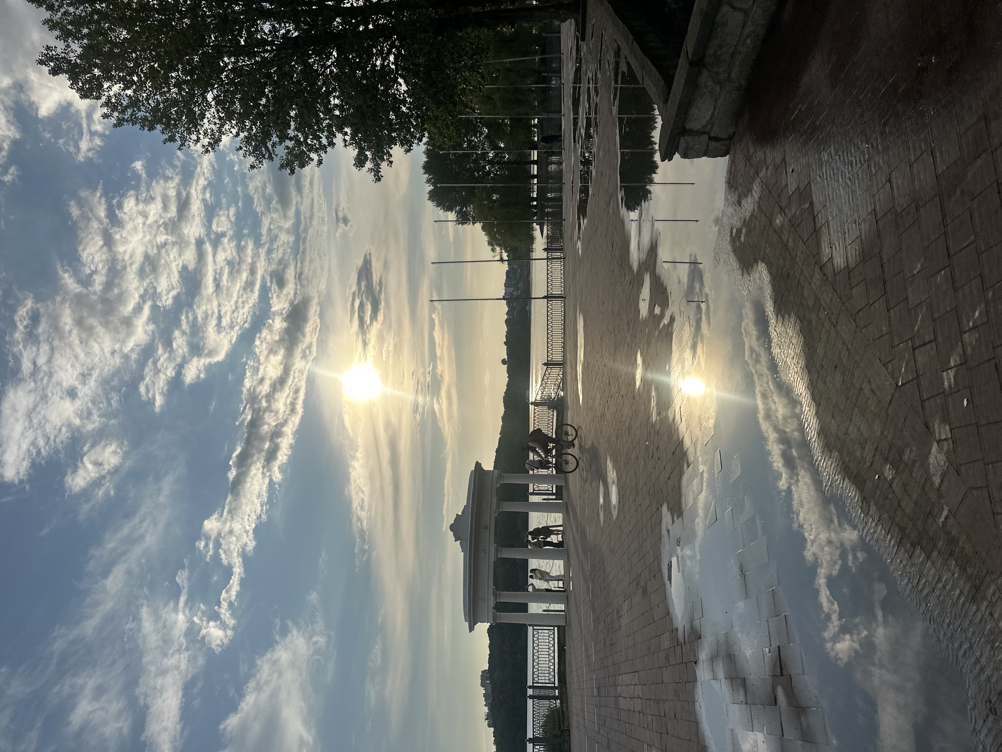 This image captures a scene at Ternopil Lake in Ternopil, Ukraine, during what appears to be late afternoon or early evening. The sky is filled with a mixture of blue and white clouds, with the sun shining brightly, creating a lens flare. In the foreground, a paved area made of reddish-brown bricks is wet, with several puddles reflecting the sky and the surrounding environment.
On the left side of the image, a white rotunda with columns stands near the lake's edge. Several people are visible near the rotunda, some standing and appearing to observe the surroundings. A person is riding a bicycle in the middle ground, moving from left to right.
The lake itself is calm, with a tree-lined shore visible on the opposite side. Further in the distance, buildings can be seen on a hillside. The right side of the image features a large tree with dark green foliage, casting a shadow on the paved area. Several tall, thin poles are also present, possibly flagpoles, in front of a line of trees. The overall impression is one of a peaceful, urban waterfront scene with elements of nature and human activity.