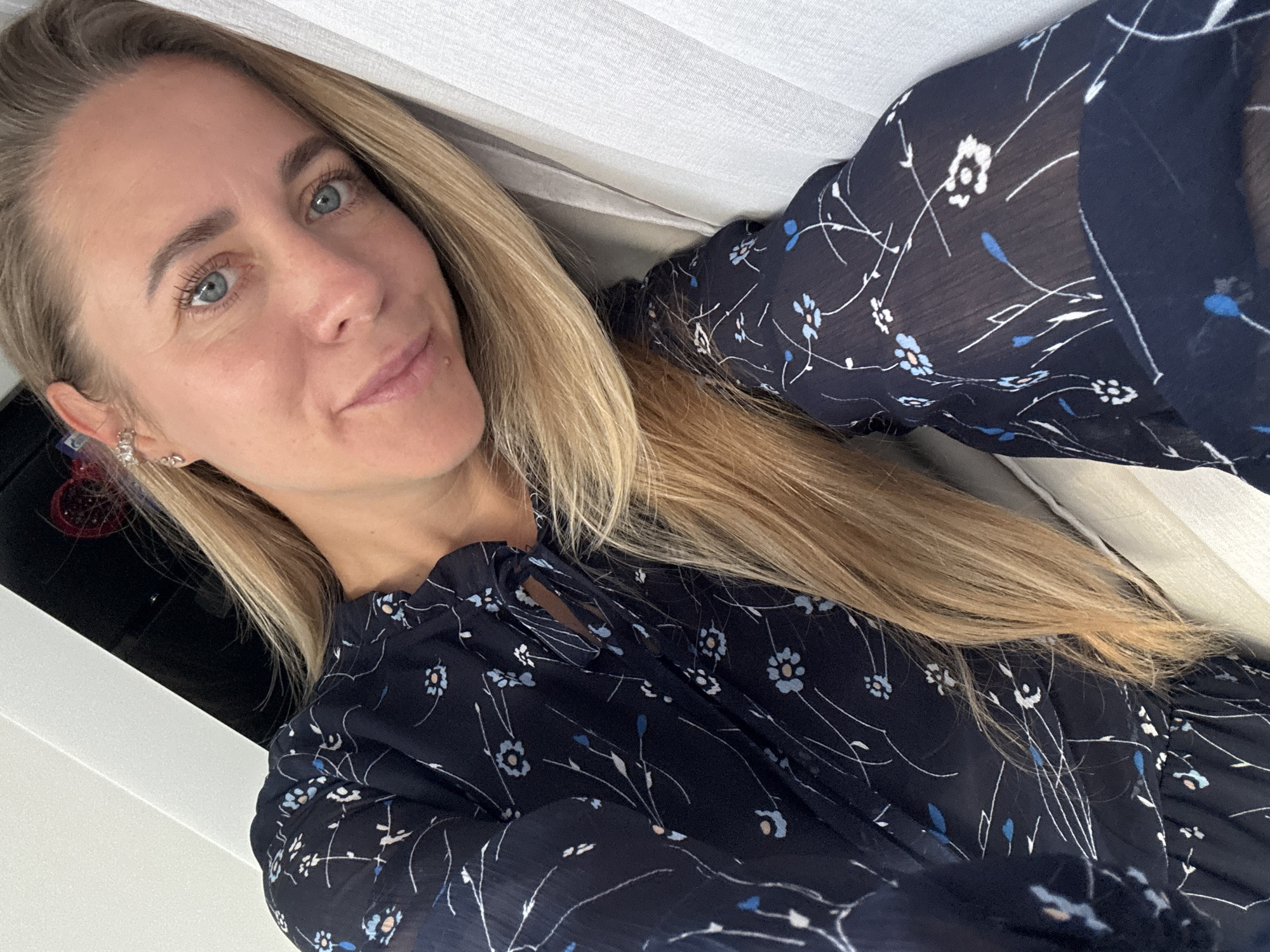 A blonde woman is leaning against a white curtain and a black surface. She is wearing a dark blue dress with a floral print. She is looking at the camera with a slight smile. Her hair is long and blonde. The image appears to be taken indoors. The location is not specified in Chaikovsky, Russia.