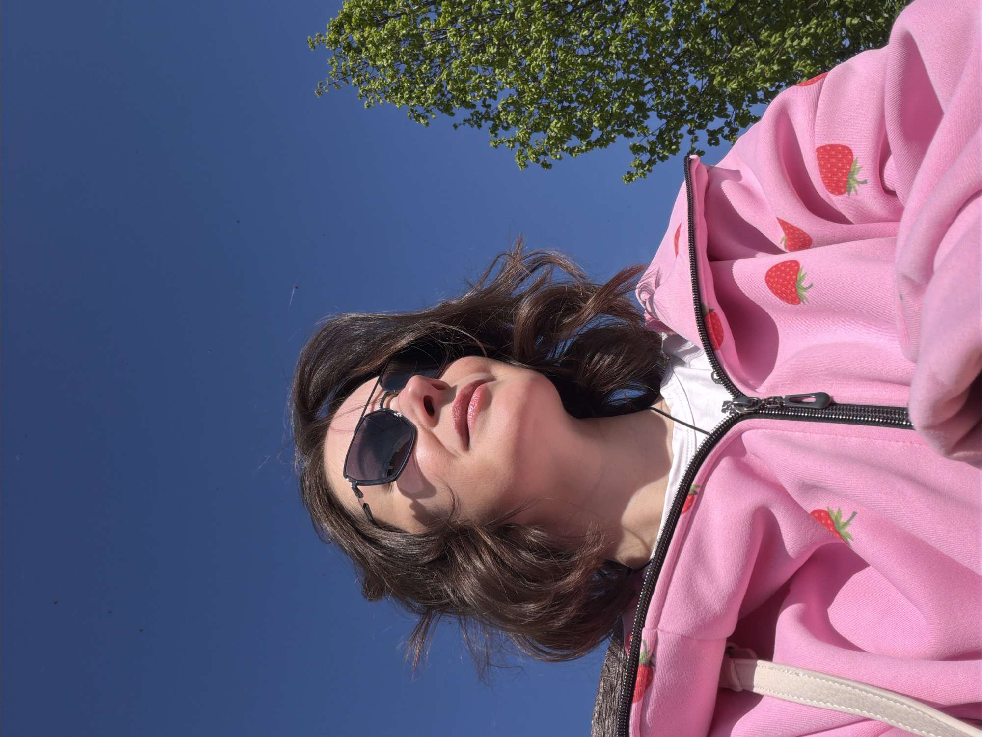 A young woman wearing sunglasses and a pink strawberry-patterned hoodie is positioned against a clear blue sky and a tree with green leaves. The photograph is taken from a low angle, looking upwards. The location is the city of Unknown, Unknown.