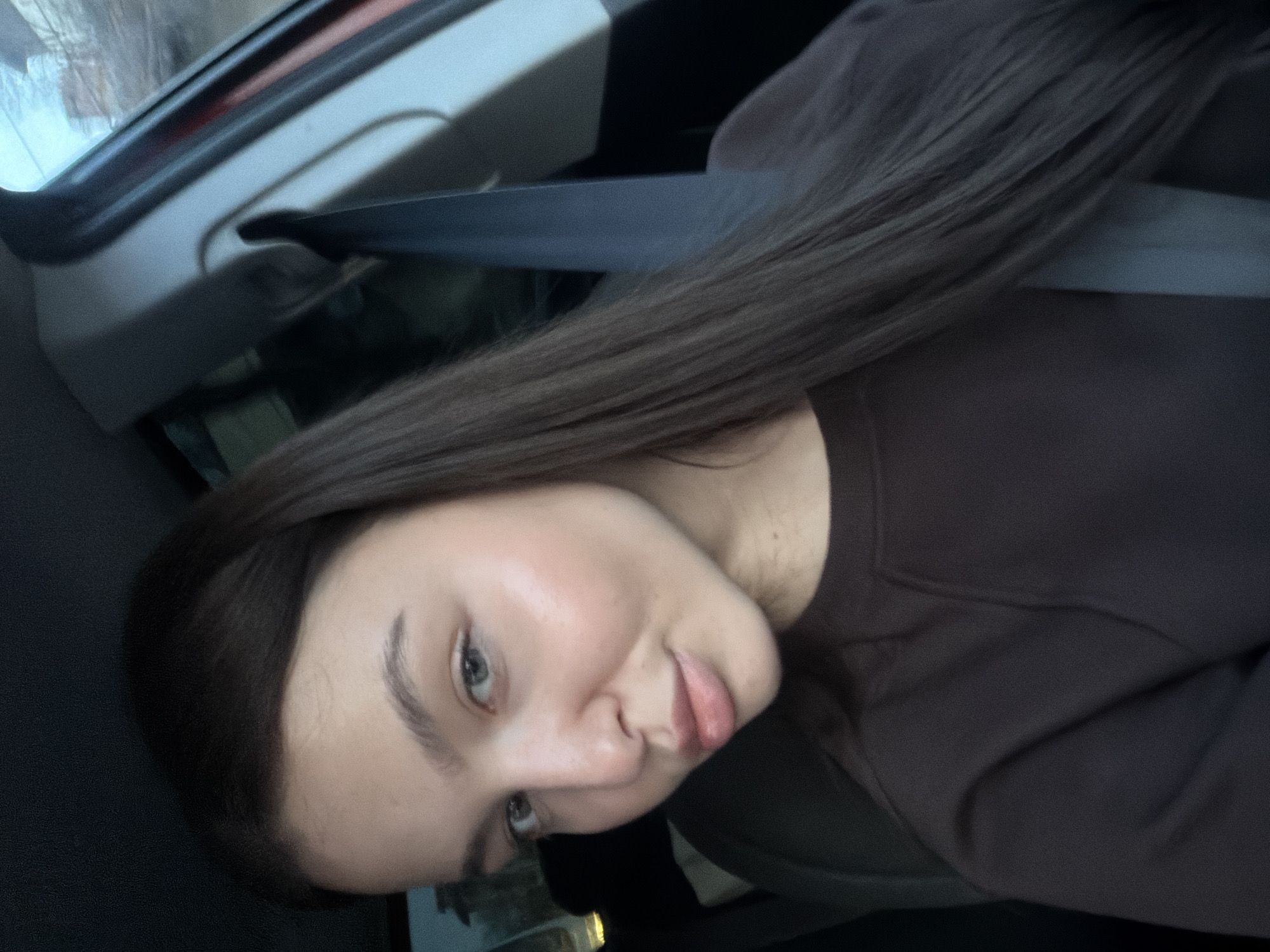A female individual, appearing in her late teens or early twenties, is depicted from the upper torso, seated within the interior of a vehicle. She possesses long, straight, dark brown hair extending over her shoulders, light-colored eyes, and her lips are slightly pursed. She wears a plain, dark brown, long-sleeved crewneck top. A dark-colored seatbelt crosses diagonally from her right shoulder across her chest. Visible vehicle interior elements include a dark headliner in the upper left, a light-colored A-pillar on the right with an attached grab handle, and a partial red trim piece along the far right edge. The exterior environment, visible through the vehicle's windows, is heavily blurred, suggesting an urban setting with indistinct structures and muted natural light. The specific location within the city of Sofia, Bulgaria, cannot be determined from the visual information provided in the image.