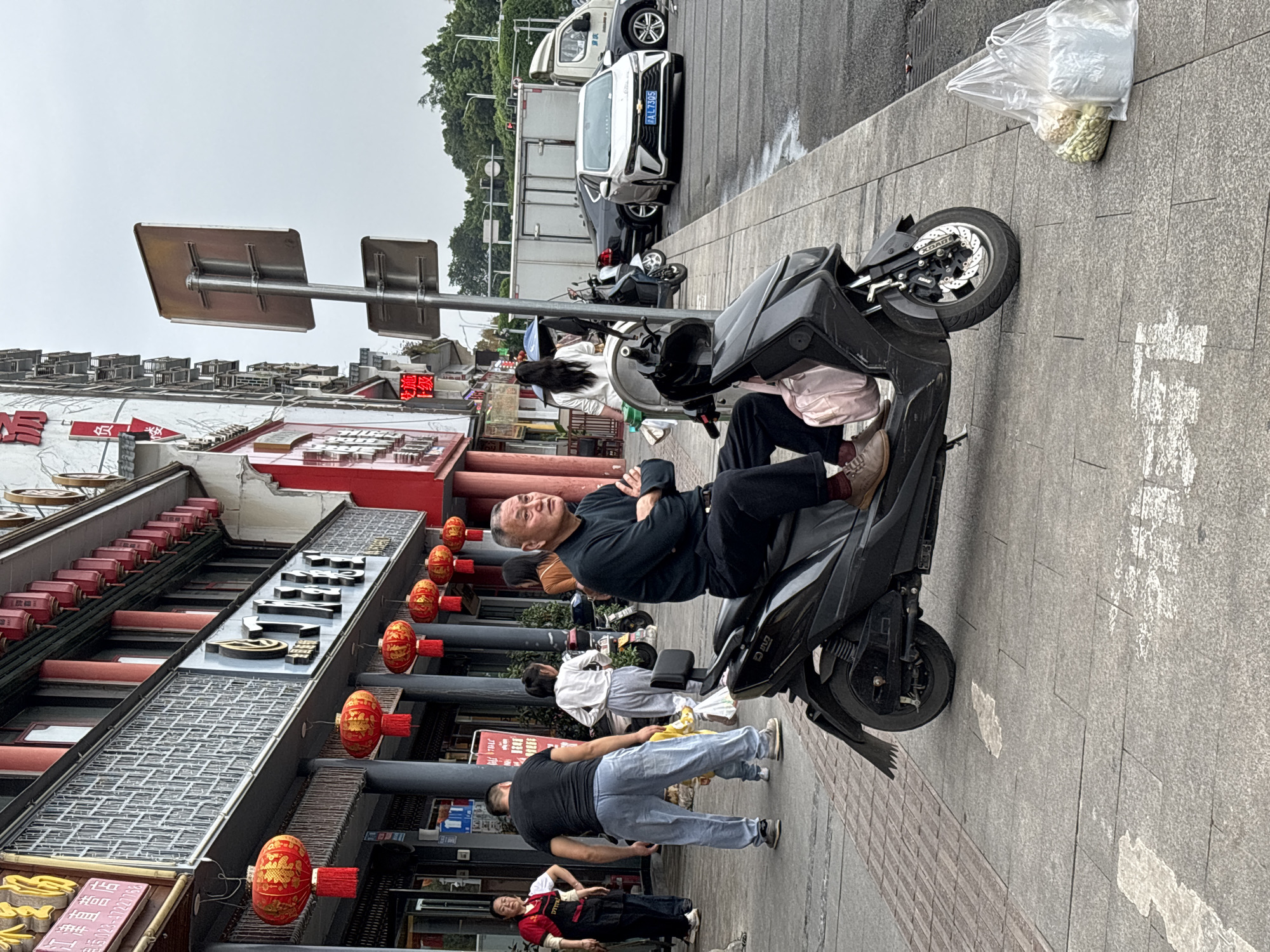 The scene depicts a sidewalk in Jiangjin, China, in front of a traditional-style building adorned with red lanterns. On the sidewalk, a man in a black shirt and black pants reclines on a black scooter with his arms crossed. Several other people are also present: one man in a black shirt and light blue pants stands nearby, and further down the sidewalk, a woman in a white shirt and light-colored pants stands next to the building. Another person in a red top and black pants is visible.
The background includes several parked vehicles. Additionally, a plastic bag containing white flowers sits on the edge of the sidewalk, and a street sign is visible in the upper left corner of the frame.
