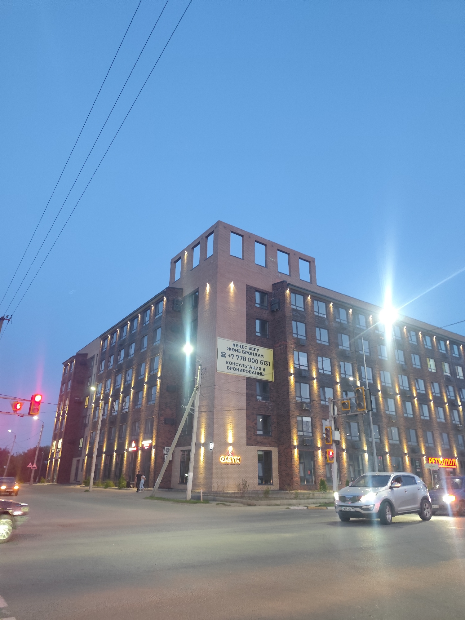 The image depicts a street intersection in Petropavl, Kazakhstan, during twilight. A multi-story modern building with brick facade and illuminated windows dominates the right side of the frame. The building has a unique architectural feature on its rooftop, with open rectangular sections. A bright advertisement banner is attached to the side of the building, featuring text in a non-Latin script and a phone number.
In the foreground, a paved road with lane markings is visible. A silver SUV is turning at the intersection, with its headlights on. To the left, a traffic light shows a red signal. Another car is partially visible in the far left of the image. A street light and power lines are also present. On the building's lower level, a sign reads "QASYM" and below it "CAFE & BARBERSHOP". Another sign on the right side of the building says "PETROPOL". Two people are standing near the building entrance. The sky is a clear blue, transitioning to darker hues of twilight.