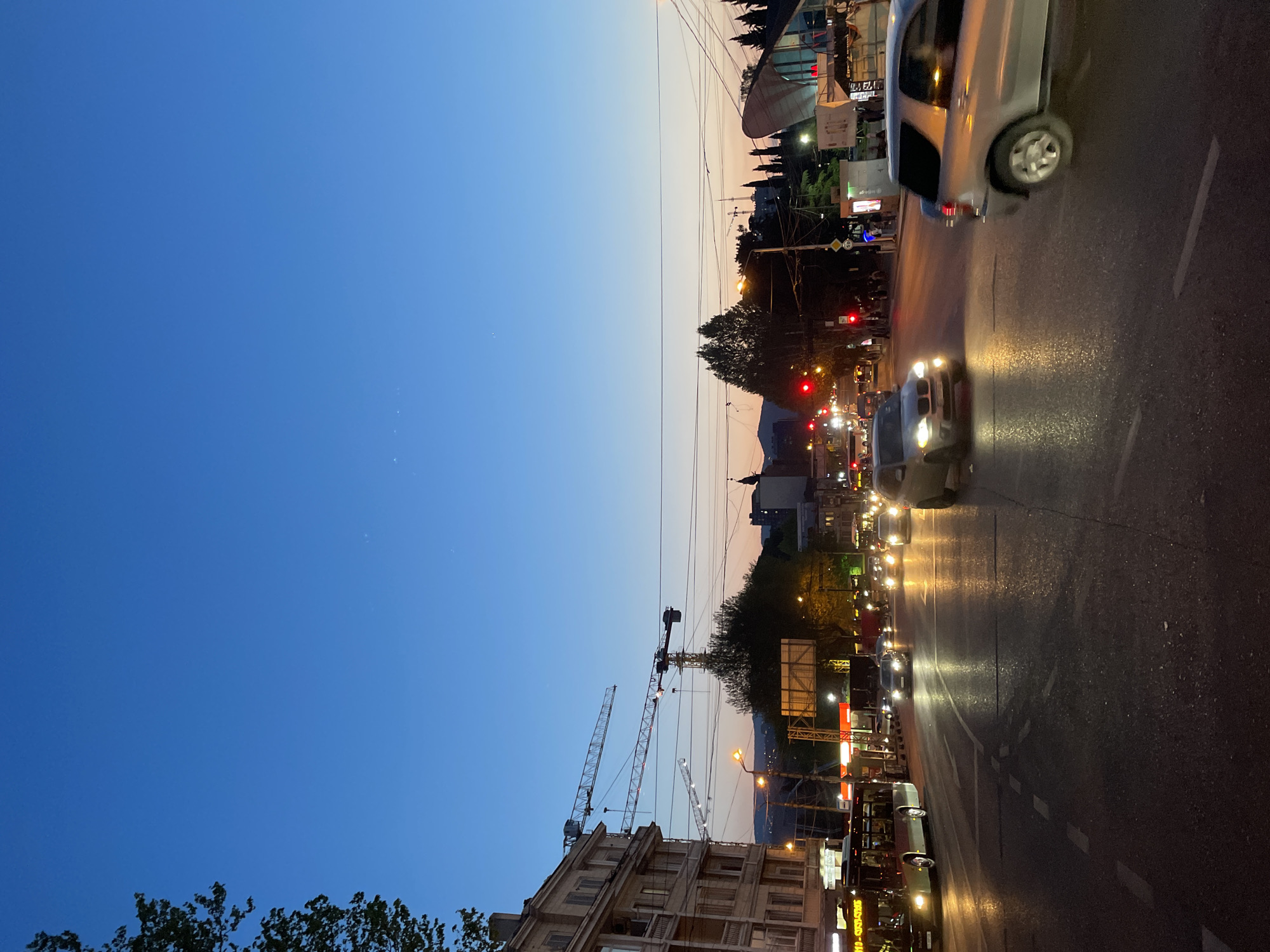 This image captures a vibrant urban street scene at twilight or early evening. The sky dominates the upper portion, transitioning from a deep blue at the zenith to a lighter, warm orange-pink gradient near the horizon, indicative of a recent sunset or impending nightfall.
The main subject is a multi-lane city street bustling with vehicle traffic. Numerous cars are visible, many with their headlights on, casting bright, elongated reflections on the wet or highly reflective asphalt road surface. A light-colored SUV is prominently featured on the right side of the road, while other vehicles, including what appears to be a white car with illuminated headlights, follow further down. Red traffic lights are visible in the mid-ground, suggesting traffic is either stopped or slowing.
Along the sides of the street, various buildings line the thoroughfare. On the left, several large construction cranes are visible towering over buildings, indicating ongoing development. Overhead utility poles and an intricate network of wires crisscross the sky, typical of a busy city environment. In the background, distant hills or mountains are silhouetted against the fading light.
Artificial light sources, including streetlights, building illuminations, and vehicle headlights, provide the primary light for the scene, creating a dynamic interplay of light and shadow. The overall impression is one of a lively, active urban setting as day transitions to night. Some signs on the buildings display text in Georgian script, reinforcing the location context of Tbilisi, Georgia.