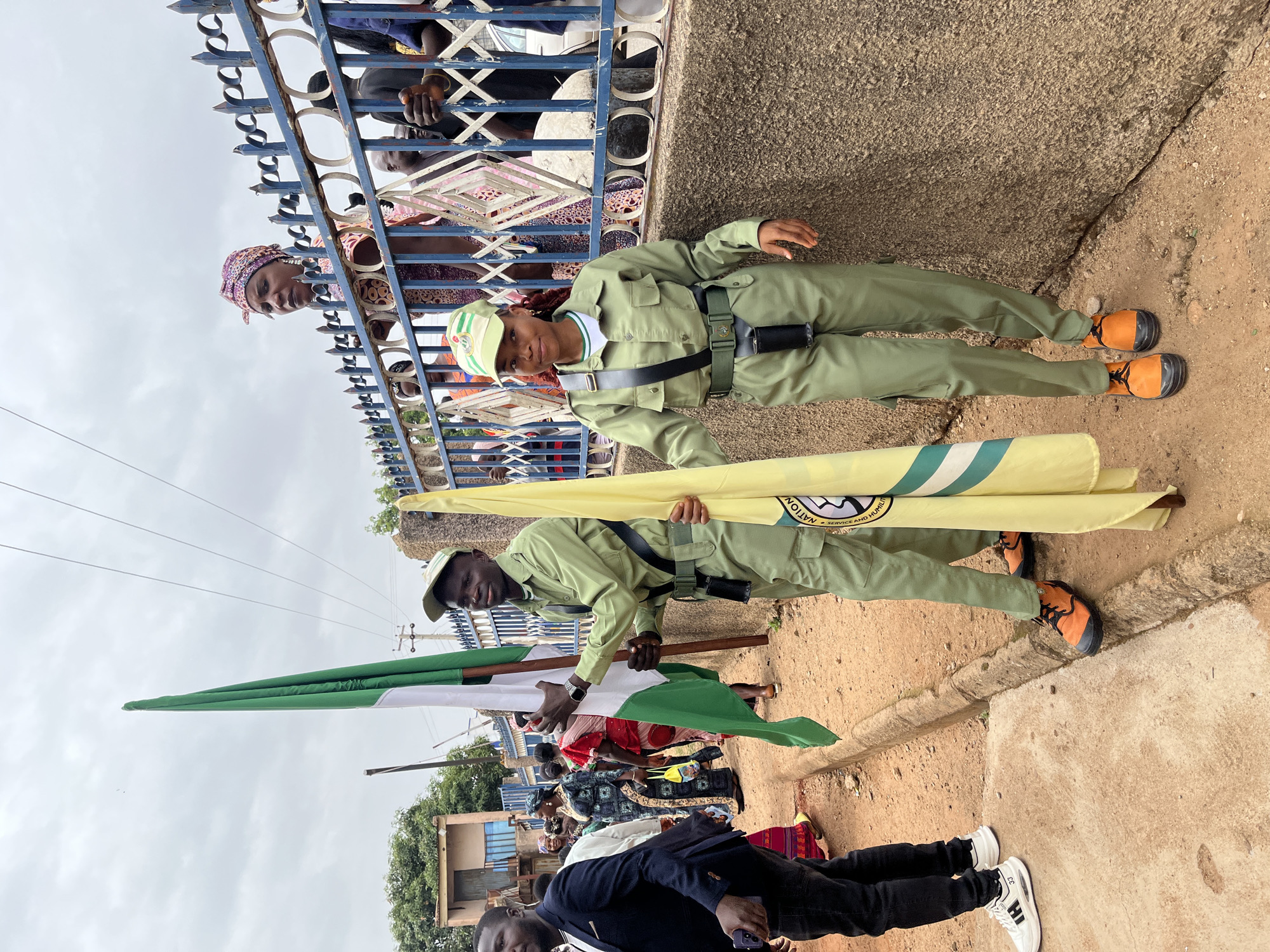 Two people dressed in khaki green National Youth Service Corps (NYSC) uniforms, complete with caps and black belts, are holding the Nigerian flag (green, white, green) and the NYSC flag (yellow with the NYSC emblem). The two people are standing by a sidewalk and a short, light-colored wall. Behind them, a blue metal fence separates them from spectators who are looking over the fence. The location is presumed to be Gura Top, Nigeria, as requested.