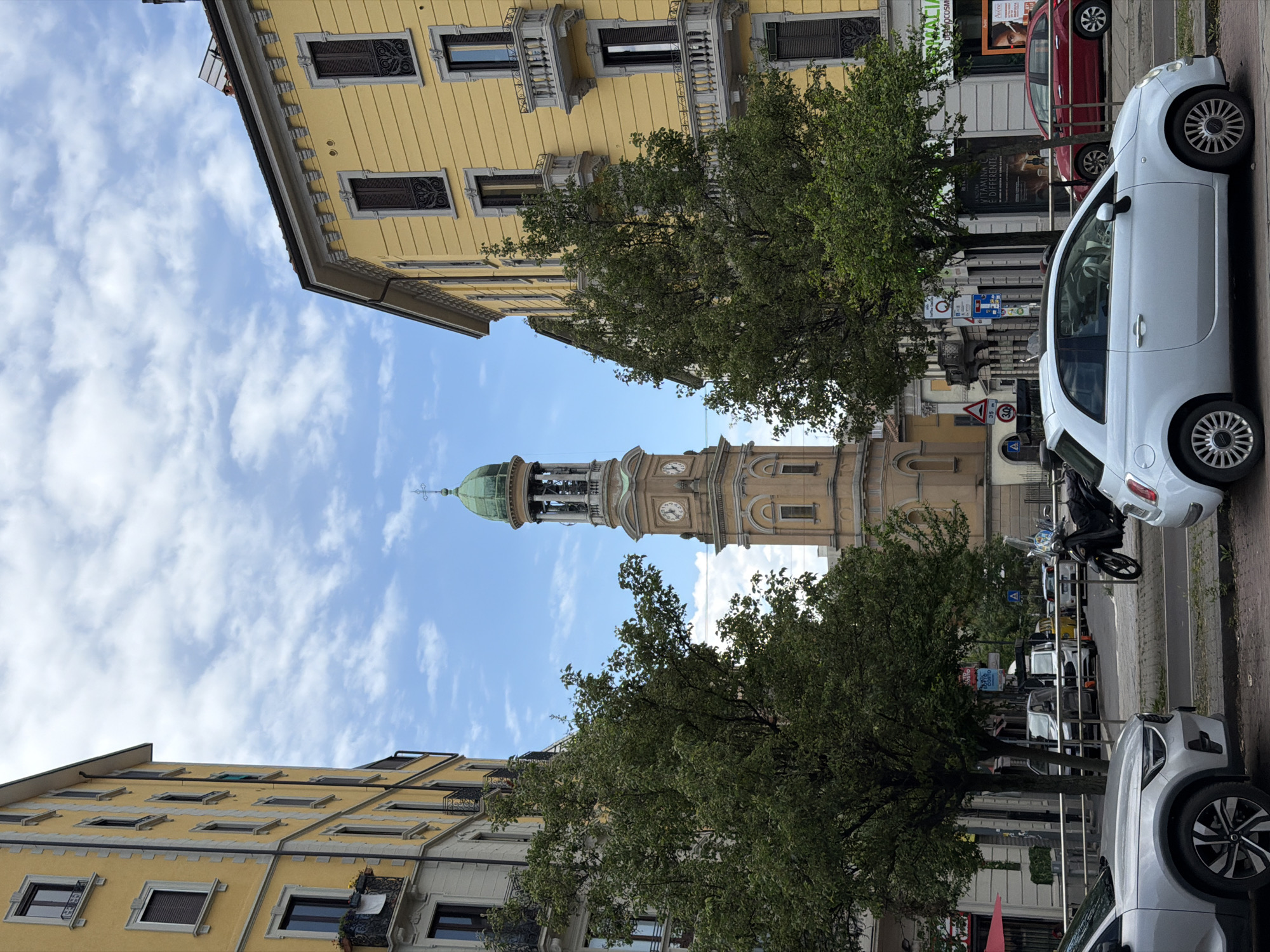 The image depicts a street scene in Milan, Italy, featuring the Torre di San Gottardo in Corte framed between yellow buildings and green trees. The tower is beige with a green dome and visible clocks on its sides. The sky is blue with scattered clouds. The street is lined with parked cars including a white Fiat 500 and a silver SUV, along with a parked motorcycle. Street signs are also visible. The buildings on either side of the street are multi-story and feature architectural details such as balconies and decorative window frames.