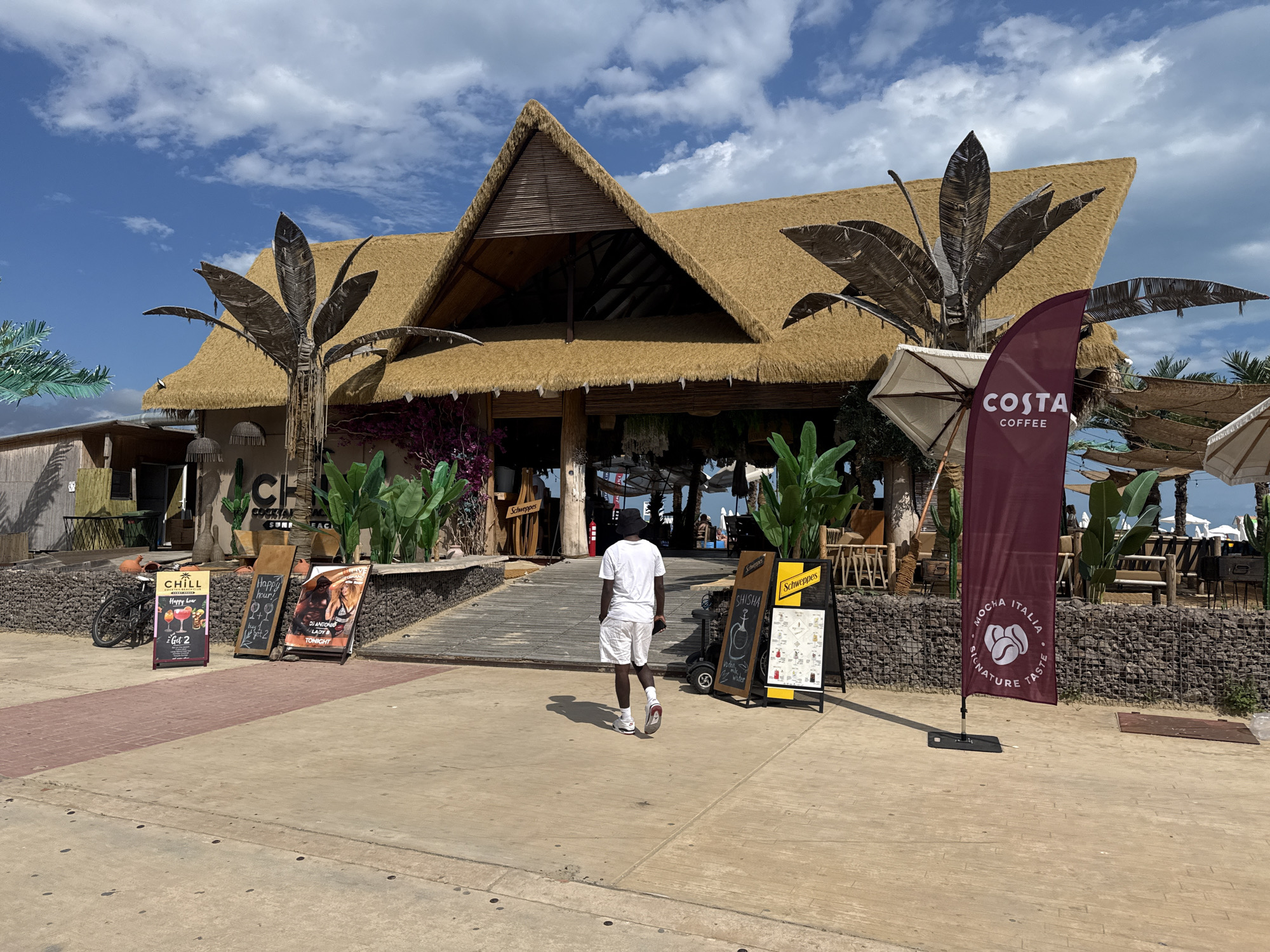 This media file captures a sunny day at a beachside establishment in Nesebar, Bulgaria. The main subject is a straw-roofed building with a prominent thatched roof, suggesting a tropical or beach-themed venue. In front of the building, a person wearing a white t-shirt and white shorts walks away from the camera towards the entrance.
Several signs are visible, advertising "CHILL COCKTAIL BAR SUNSET," "Happy Hour," and "SHISHA." One sign promotes "DJ ANTONIO & LADY B TONIGHT." A prominent maroon flag displays "COSTA COFFEE" and "MOCHA ITALIA SIGNATURE TASTE." Other objects include umbrellas, potted plants, decorative palm fronds made of wood, and a bicycle leaning against a wall. The ground is paved with concrete and brick.
The weather appears to be clear and bright with scattered clouds in a blue sky, indicating daytime. The overall atmosphere is relaxed and inviting, typical of a beachfront bar or restaurant during operating hours. The presence of the Costa Coffee flag suggests a potential partnership or offering. The setting is clearly a public outdoor space, likely a promenade or walkway leading to the establishment.