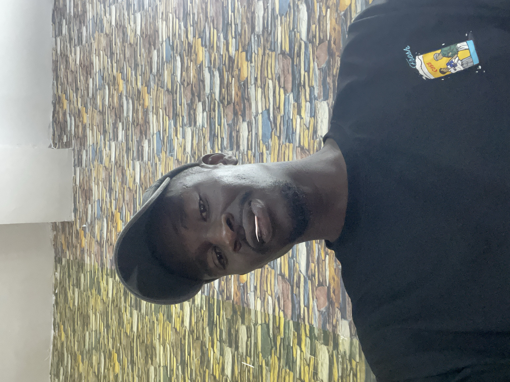A portrait shows a Black man with a dark beard, wearing a dark baseball cap and a black t-shirt with a cartoon design on the upper right corner. He is turned slightly, looking at the camera with a slight smile and a piece of gum sticking out of his mouth. The backdrop is a wall with textured, multi-colored stone-like cladding. There is no specific information about Jos, Nigeria, or details to confirm the location.
