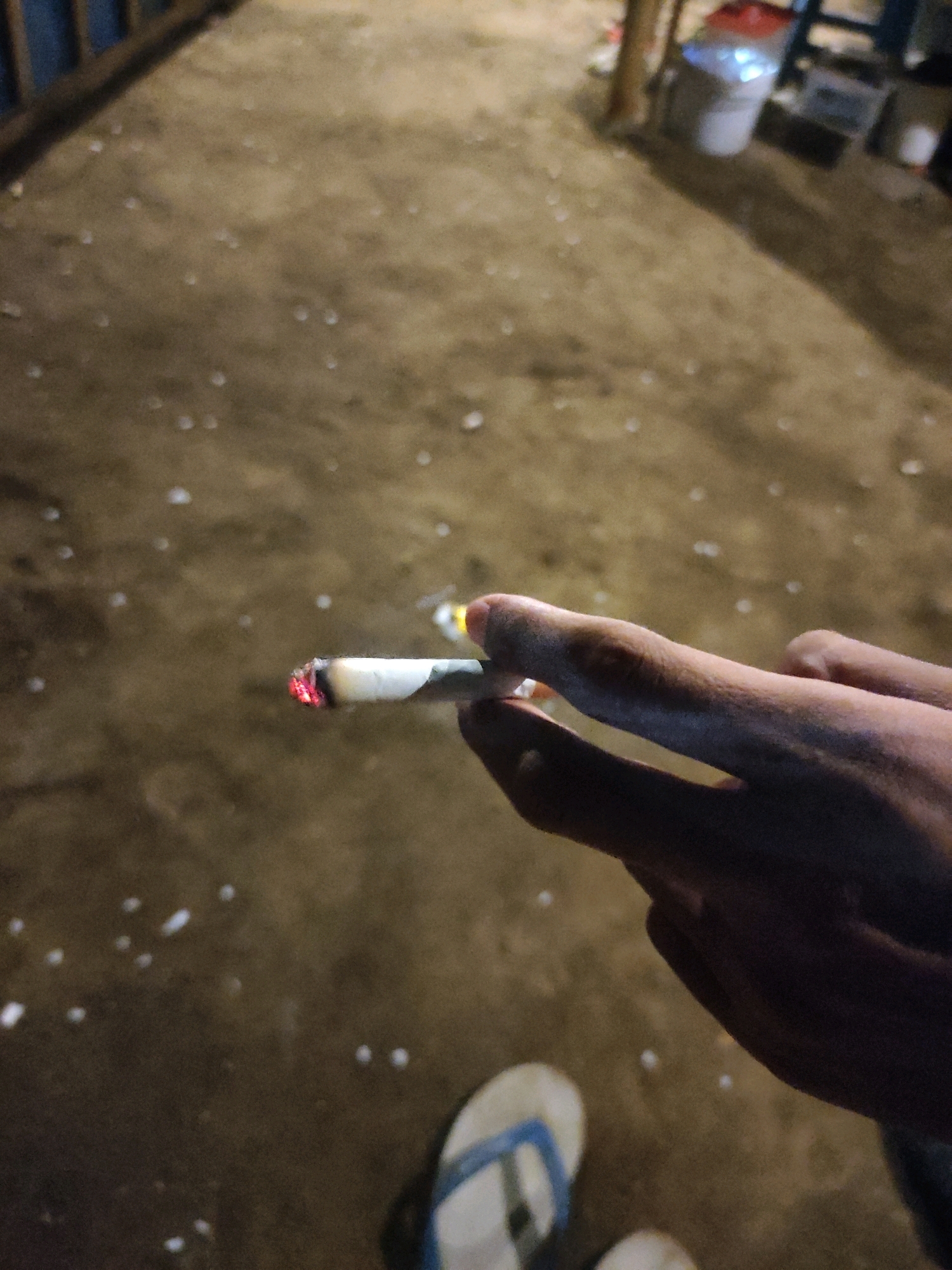 A close-up shot focuses on a hand holding a lit cigarette. The cigarette is partially consumed, with the burning end glowing red. The hand appears to be bare, with fingers gently cupping the cigarette. The background is a dirt surface, possibly a pathway or ground, with scattered white debris. The lighting suggests it is nighttime or a dimly lit indoor environment. The image does not provide any specific clues about the location within Tiudan, Indonesia.