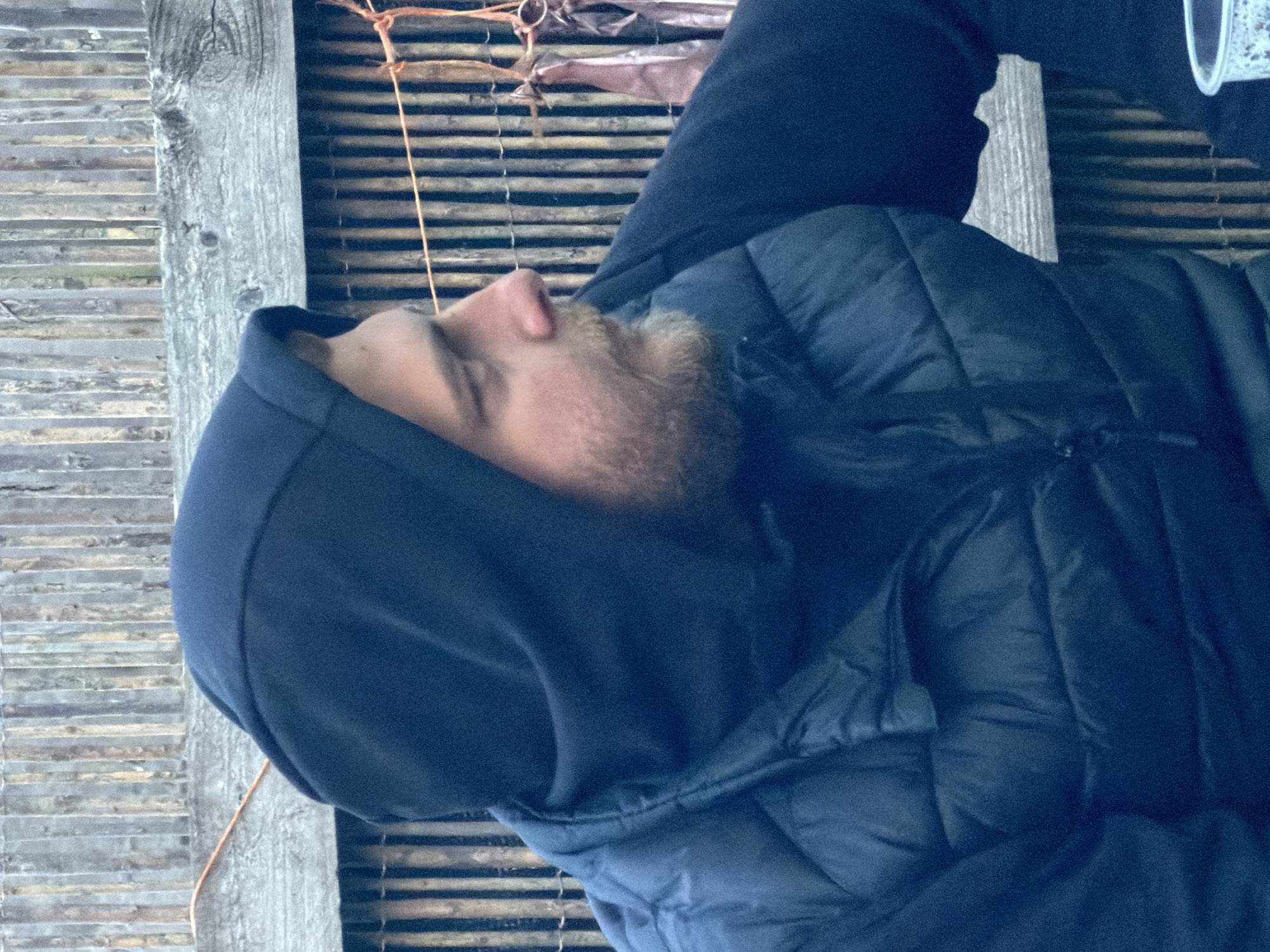 A male individual is depicted in a right-facing profile, wearing a navy blue hooded sweatshirt with the hood up and a matching navy blue quilted puffer vest over it. He has a short, light-colored beard or stubble. His gaze is directed forward and slightly downward.
The background consists of a rustic wooden fence or wall made of numerous vertical wooden poles or slats of varying widths and weathered tones. A prominent horizontal wooden plank or beam runs across the mid-ground. A thin, orange-brown rope is strung horizontally across the fence, from which several elongated, somewhat indistinct pinkish-brown items, possibly fabric or dried organic material, are suspended by smaller strings.
In the bottom right foreground, a portion of a white plastic cup, revealing some internal texture or residue, is visible. The lighting is natural and even, suggesting an outdoor or semi-outdoor, informal setting. The location in the city of Maidstone, United Kingdom, cannot be independently verified from the visual information present in the image.