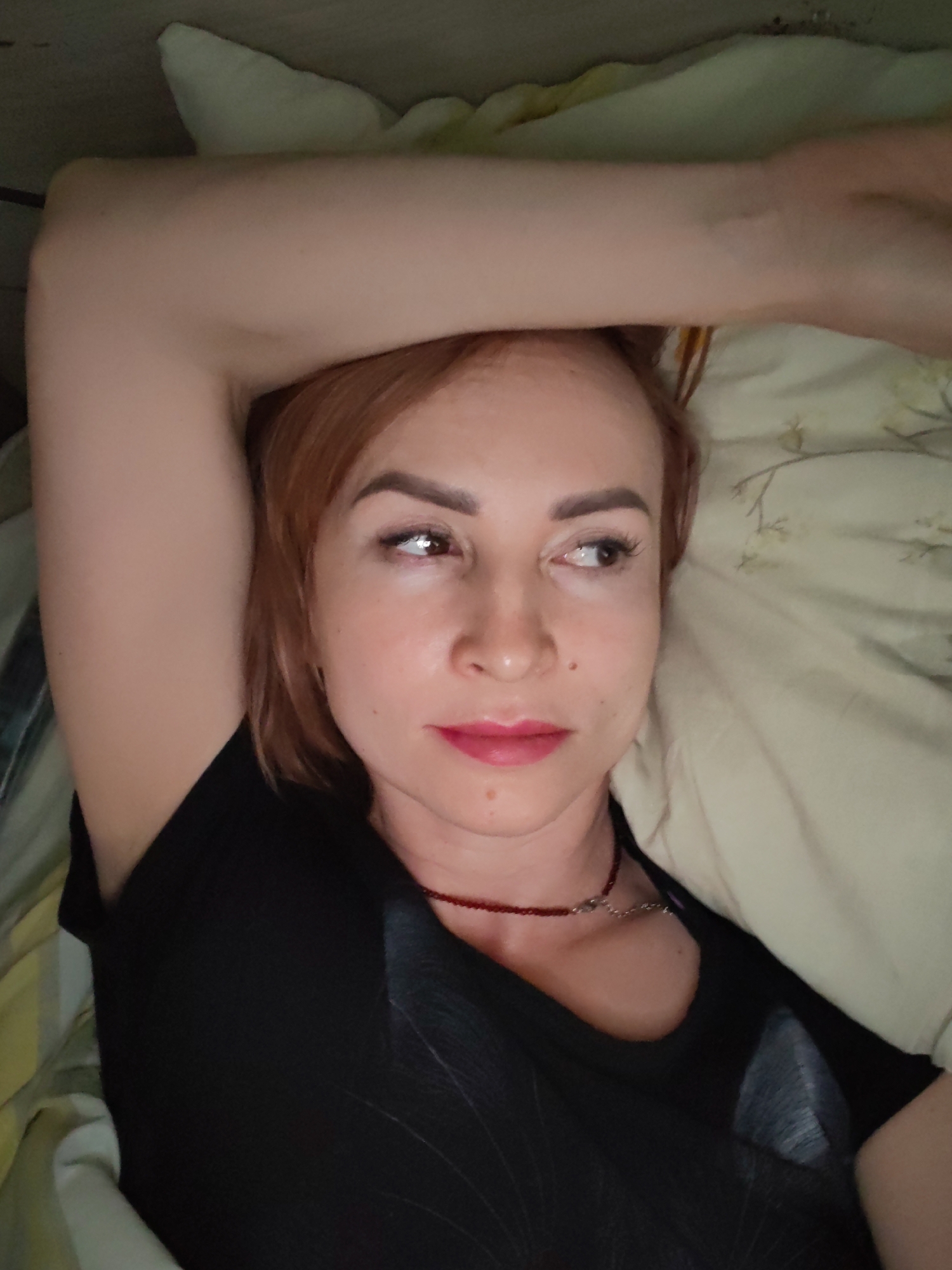 A woman with auburn hair is lying on a bed, possibly in a residential setting, though a specific location in Chaikovsky, Russia is not discernible from the image. She is wearing a black shirt and a red beaded necklace. Her right arm is raised above her head, resting on a pillow. Her gaze is directed to the side. The bed is covered with patterned sheets and pillowcases.