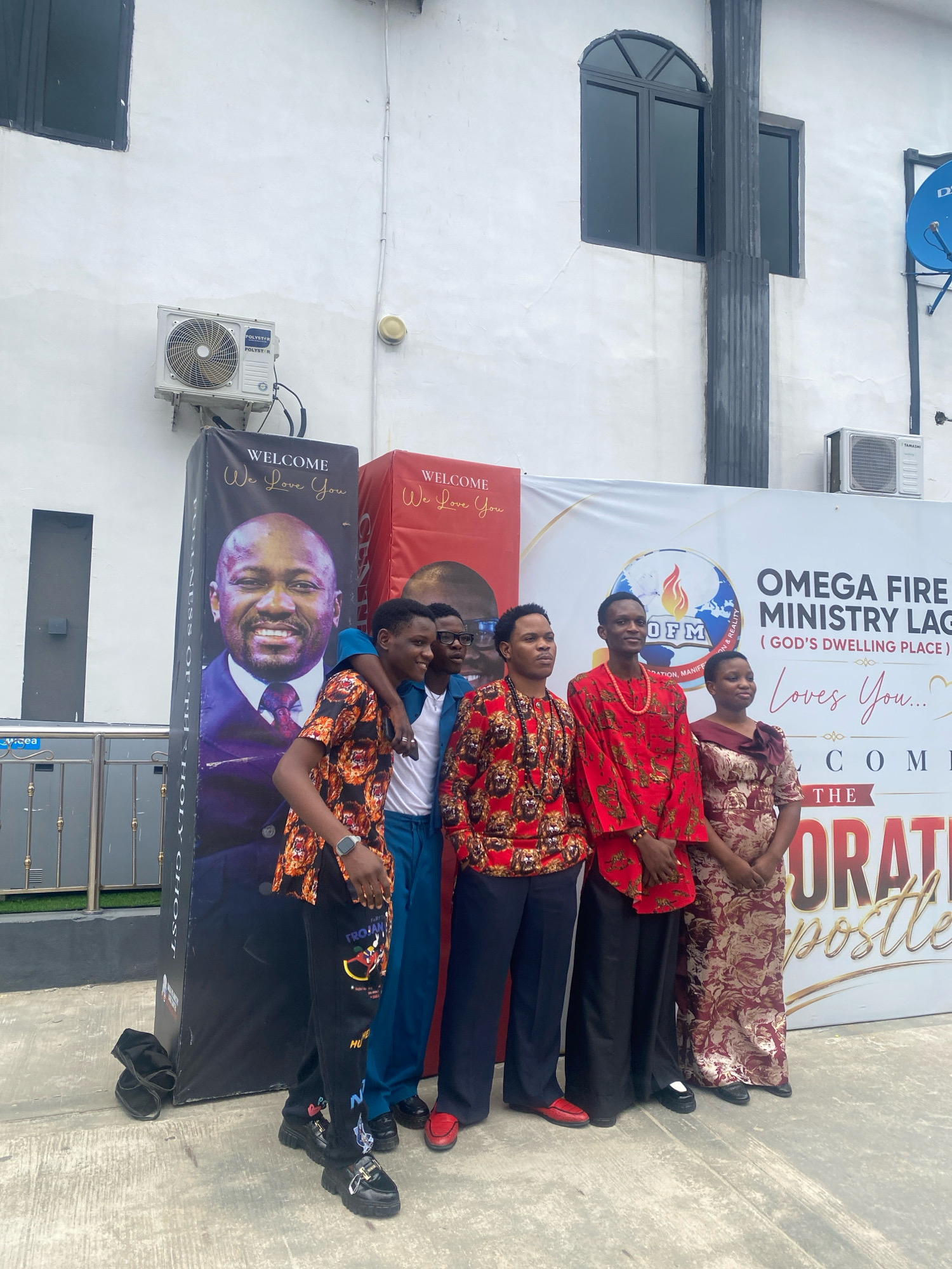 In Ado, Nigeria, a group of five individuals is posing for a photograph outside a building. The group consists of four men and one woman. The man on the far left is wearing a black shirt with an orange and black tiger-like print, black pants with colorful graphic designs, and black chunky platform shoes. He has a watch on his left wrist. Next to him is a man in a blue shirt and blue pants, with black shoes. To his right is a man wearing a red printed shirt with a gold beaded necklace and black pants. He is followed by a man in a red printed shirt and black pants. The woman on the far right is wearing a patterned maroon and gold dress. Behind the group, there is a large banner that says "WELCOME We Love You" and features a portrait of a man. Another banner behind them reads "OMEGA FIRE MINISTRY LAGOS (GOD'S DWELLING PLACE) Loves You... WELCOME THE". The building has a white facade with a dark gray column and windows. An air conditioning unit is mounted on the wall.