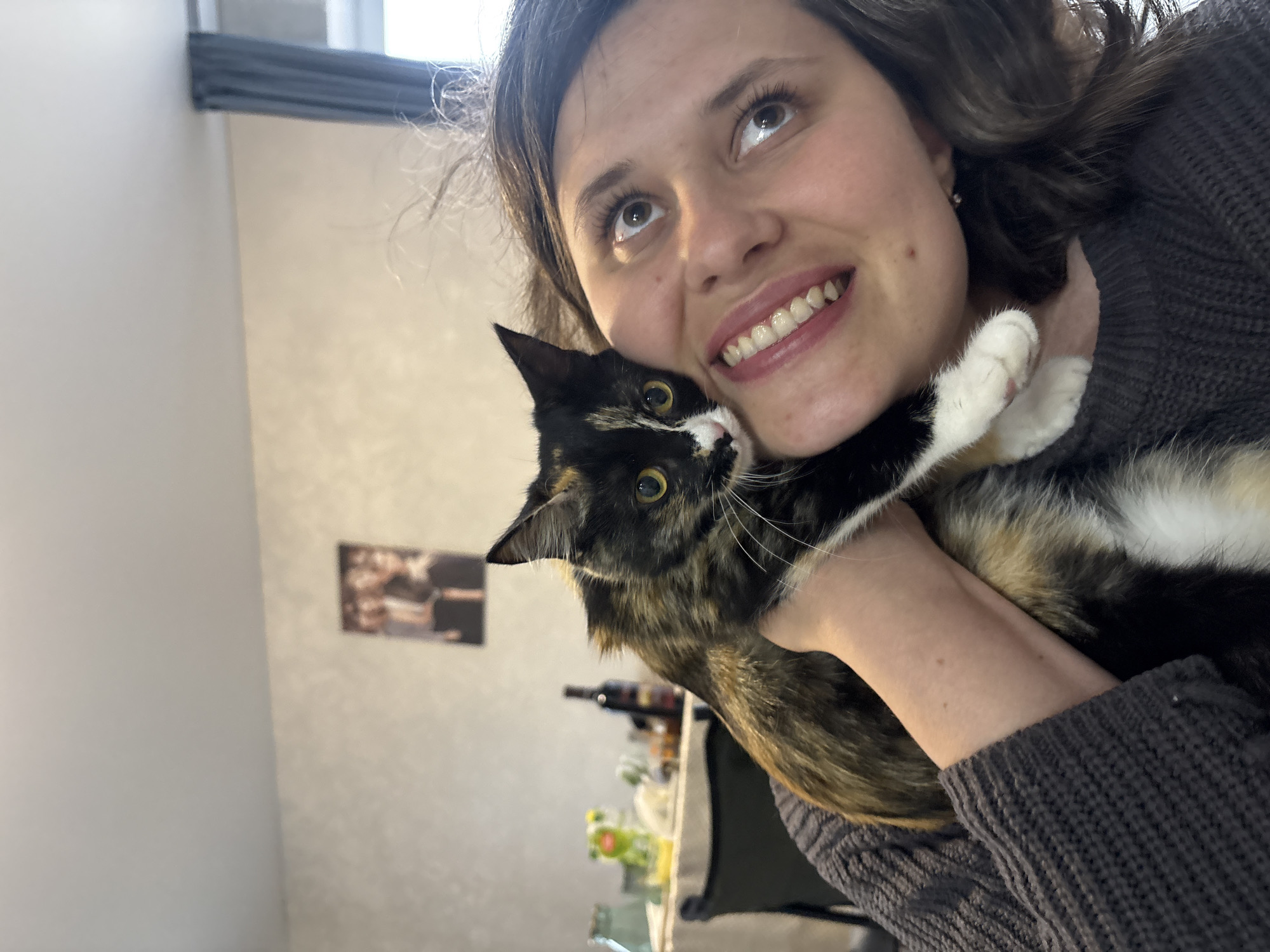 A smiling woman with brown hair and brown eyes holds a tortoiseshell and white cat. The woman is wearing a gray sweater. Behind the woman and cat, there is a light-colored wall with a picture hanging on it. There is a window at the top of the frame. In the background, on a surface, there are various objects including a wine bottle, a chair, and food items. The city of Прикамский, Russia cannot be inferred from the image.