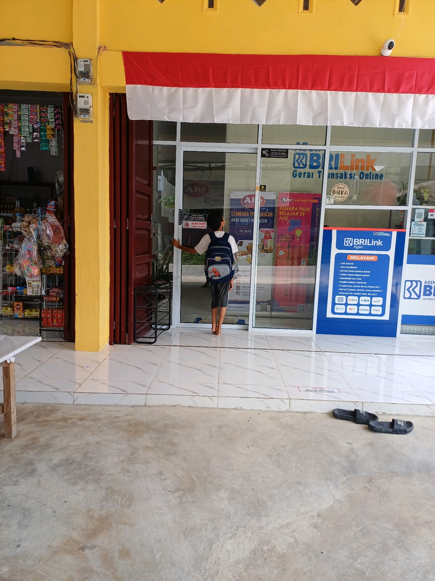 This is a street-level, medium-shot photograph taken outdoors in Dumai City, Indonesia. The scene depicts the exterior of a small shop, likely a convenience store or a small business operating as a financial agent. The building's facade is painted in a bright yellow, with a red and white Indonesian flag draped across the top of the entrance.
In the foreground, the ground is a mixture of concrete and white tiled flooring. Two black flip-flops are placed on the concrete to the right of the frame. A wooden table leg is visible in the lower left corner.
The shop entrance is a glass door, revealing the interior. A young person, seen from the back, is standing at the entrance, partially inside the shop. They are wearing a dark backpack and dark shorts, and appear to be barefoot. Inside the shop, shelves stocked with various goods, predominantly packaged snacks and beverages, are visible.
The glass door also displays several signs and stickers. Notably, there is a blue sign with white text that reads "BRI Link Agen MELAYANI" and lists various financial services offered. Other signs in Indonesian, some with logos like "Ahe" and "BRI," are also present on the glass.
To the left of the main entrance, a separate section of the building, painted yellow, appears to be another entrance or part of the same shop, with shelves of goods visible behind a glass pane. Electrical meters are mounted on the yellow wall above this section.
The overall impression is that of a bustling local business in a residential or commercial area of Dumai City.